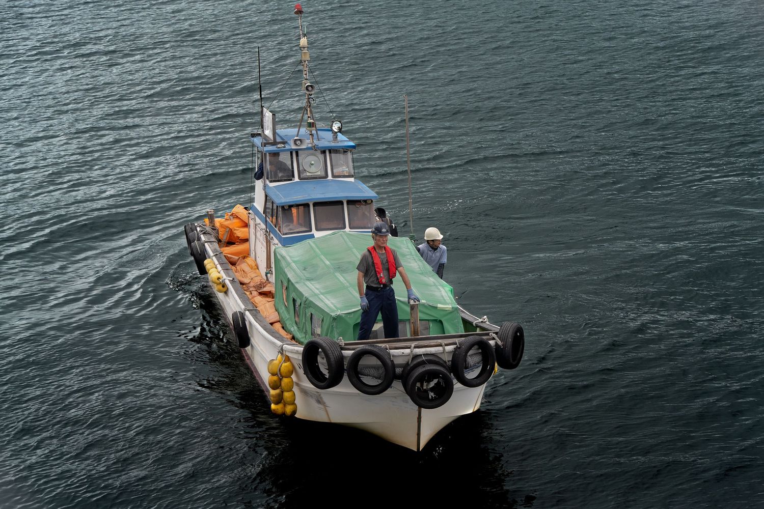 Small fishing boat with green tarp and tires on calm water, with crew member standing on deck wearing a hat.