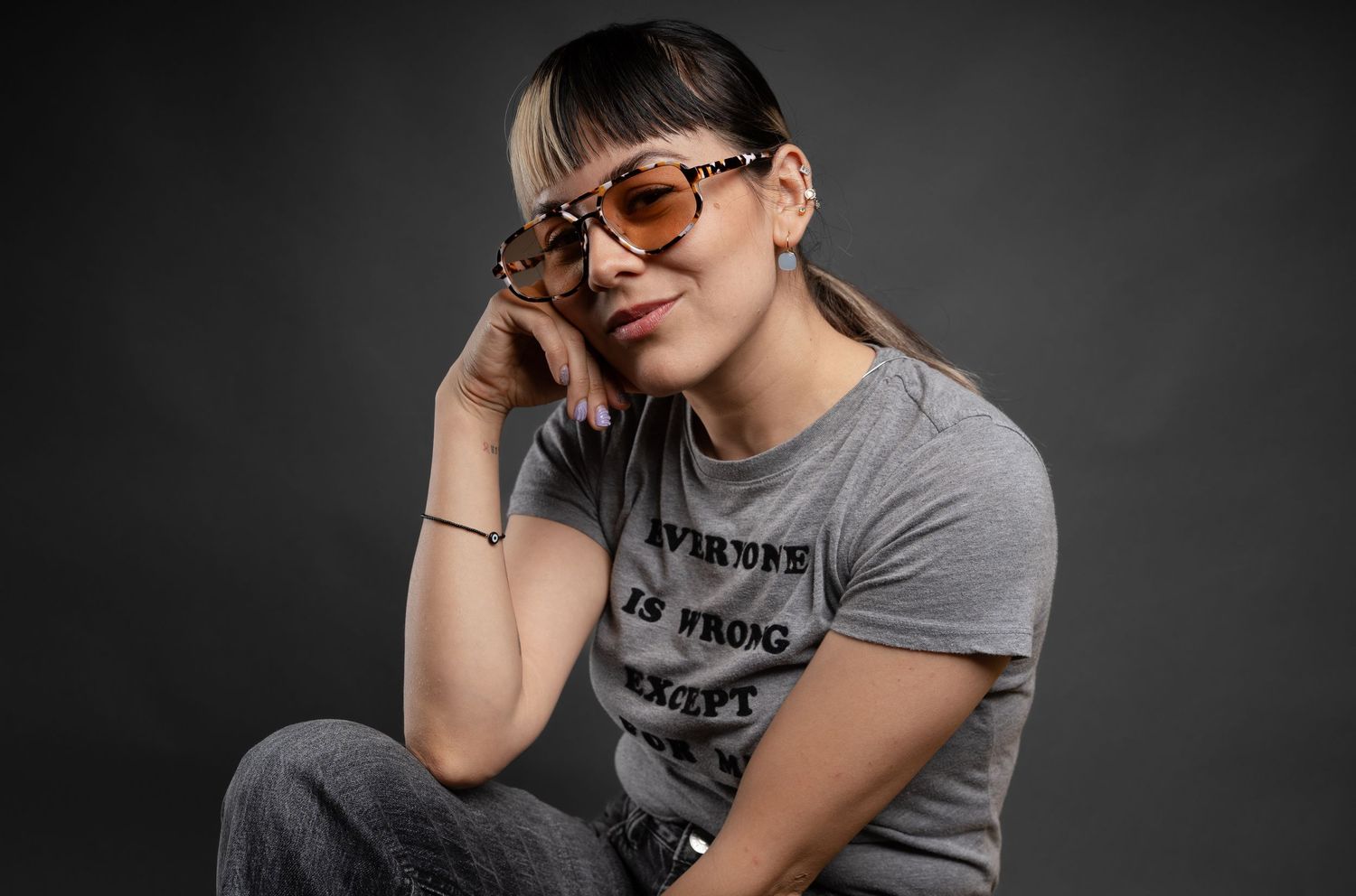 Person in glasses and gray t-shirt posing expressively against dark studio background.