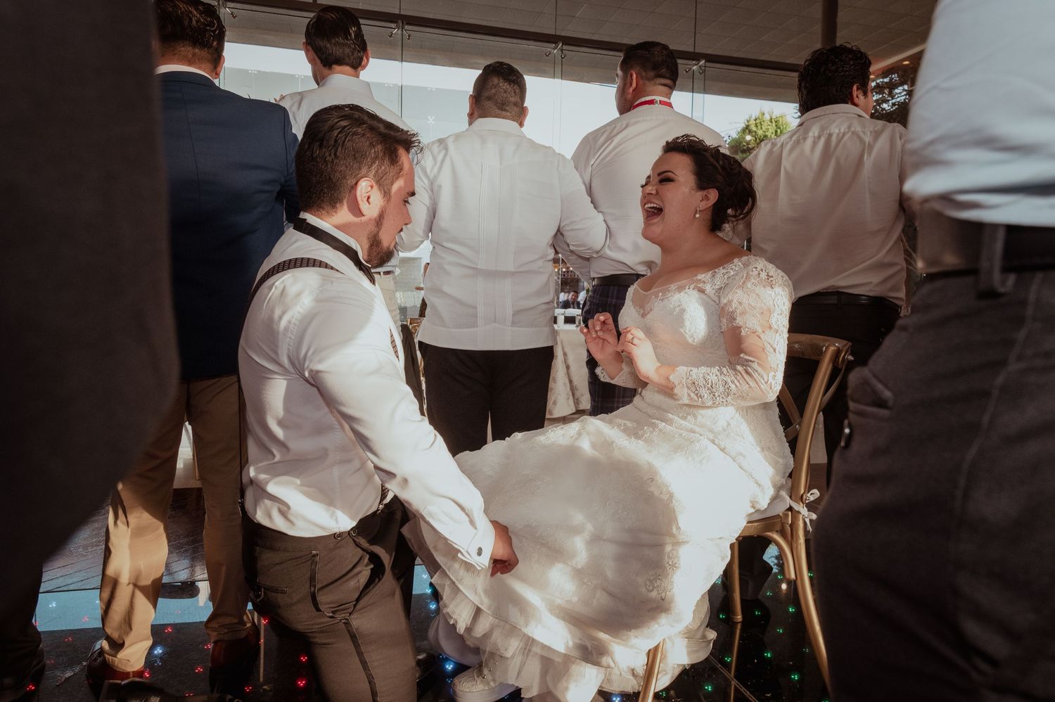 Bride in white lace gown laughing joyfully with groom in suspenders at wedding reception.