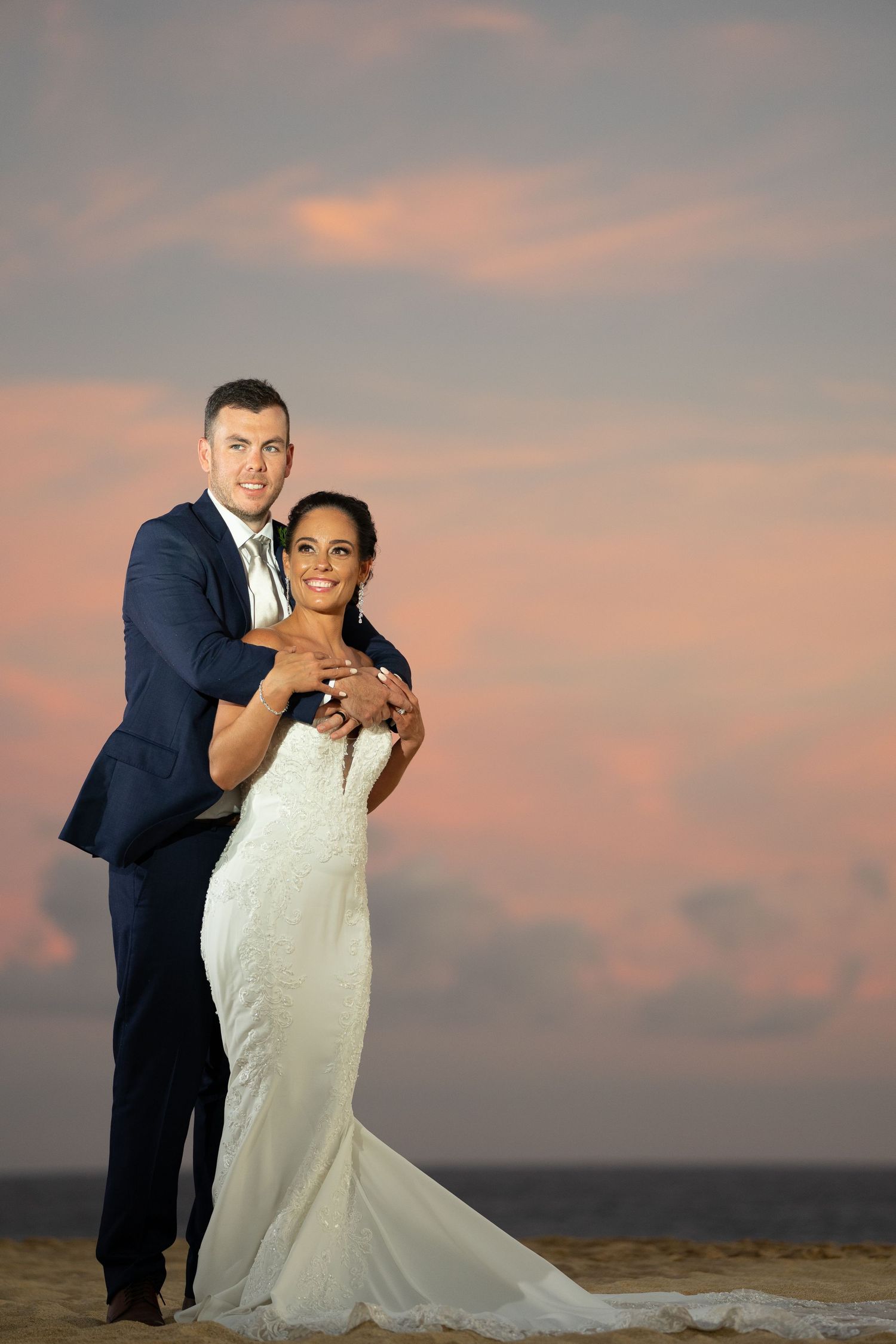 Bride and groom embracing on beach at sunset, wearing white gown and navy suit, smiling at camera.