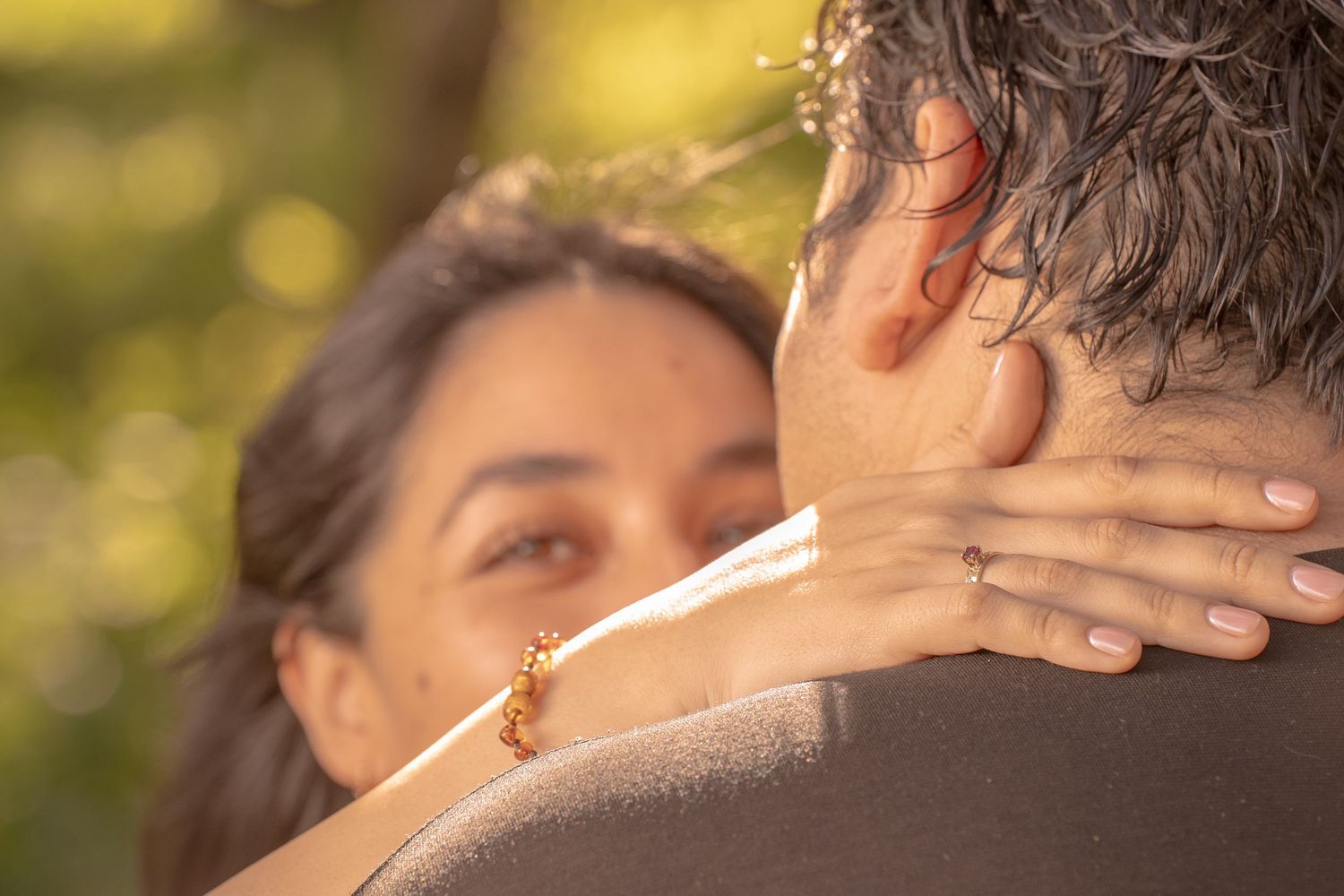 A tender embrace captured from behind with hands clasped around neck in soft sunlight.
