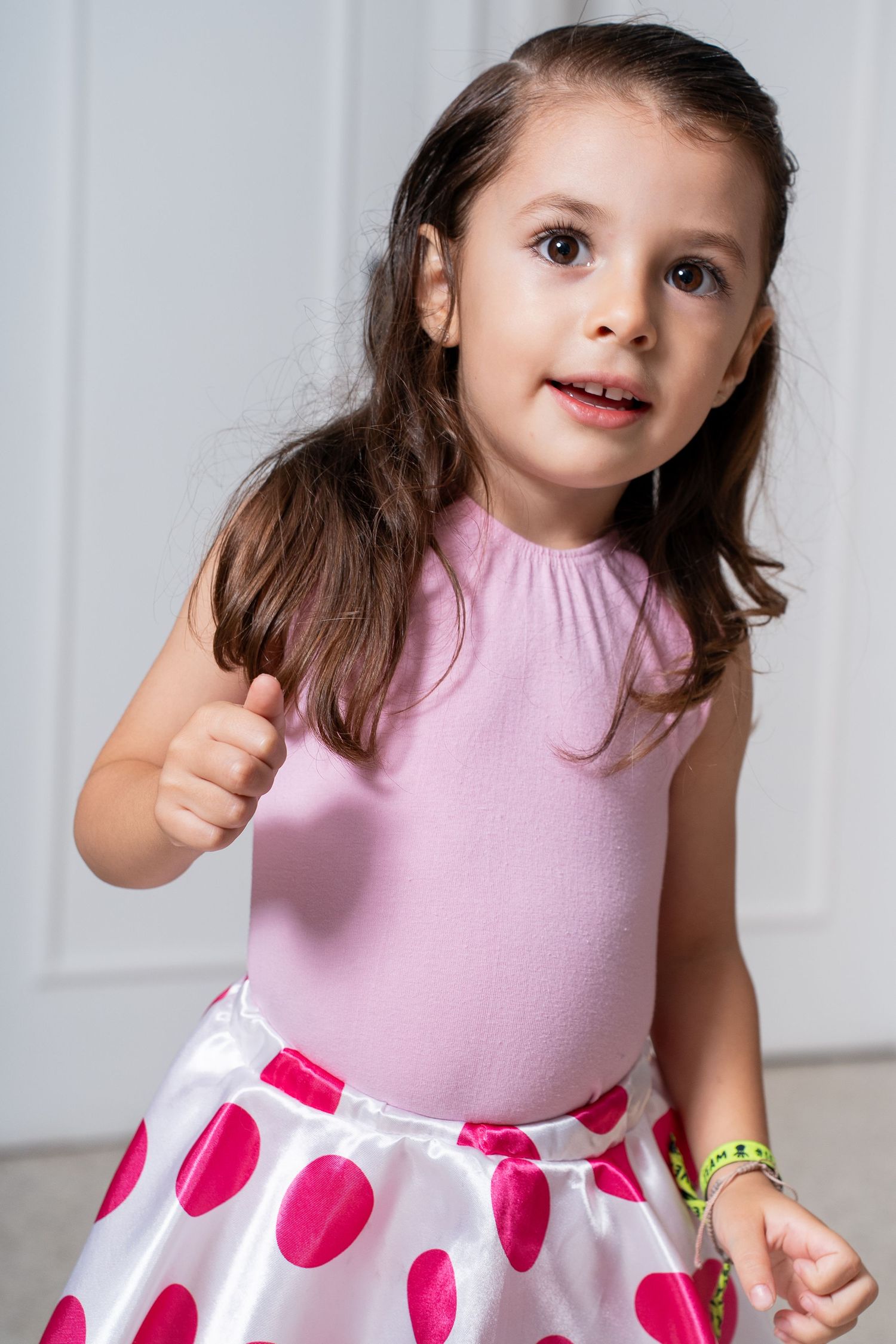 Young girl in a pink and polka dot dress posing playfully against a white background.
