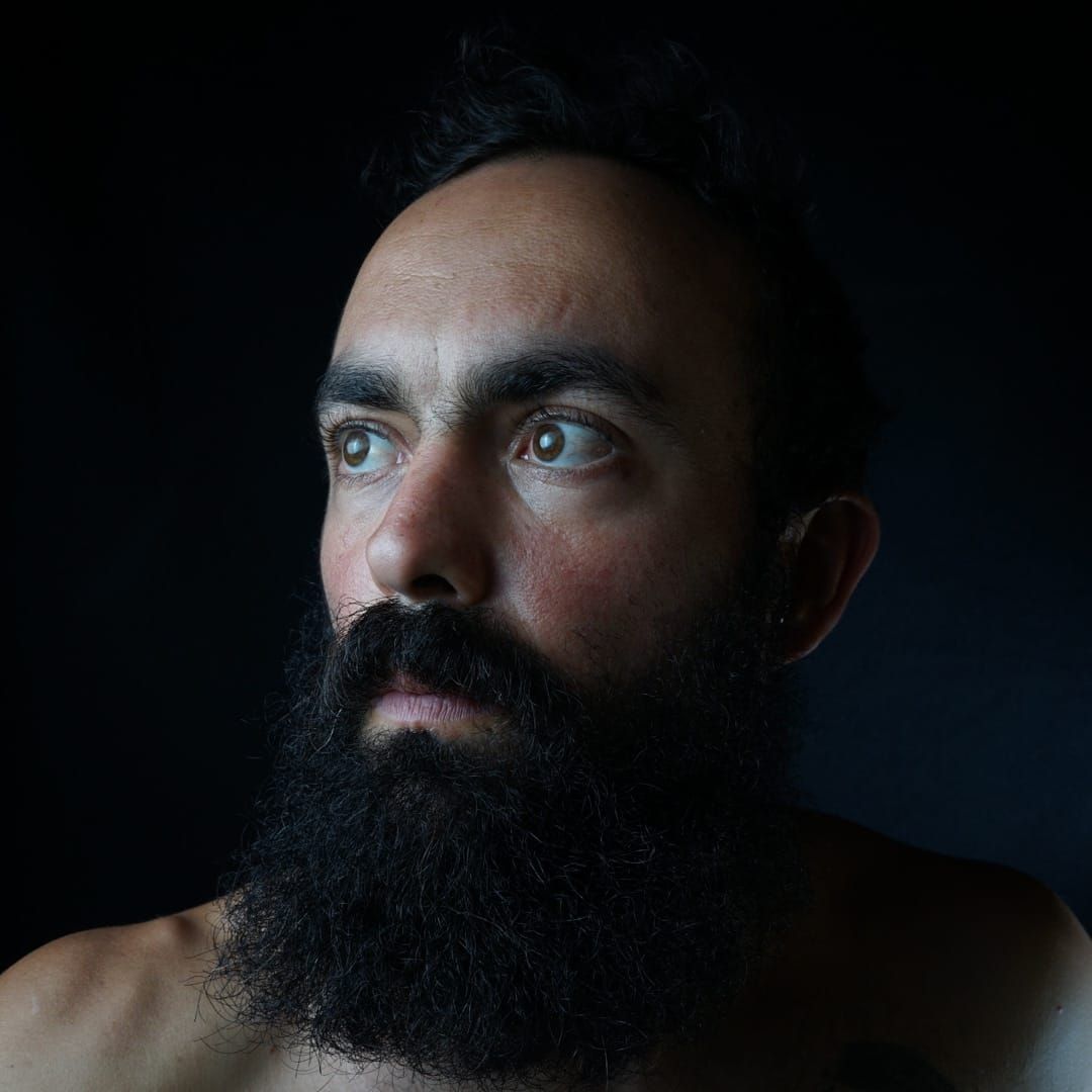 Dramatic side-lit portrait against dark background showcasing a full dark beard and intense gaze.