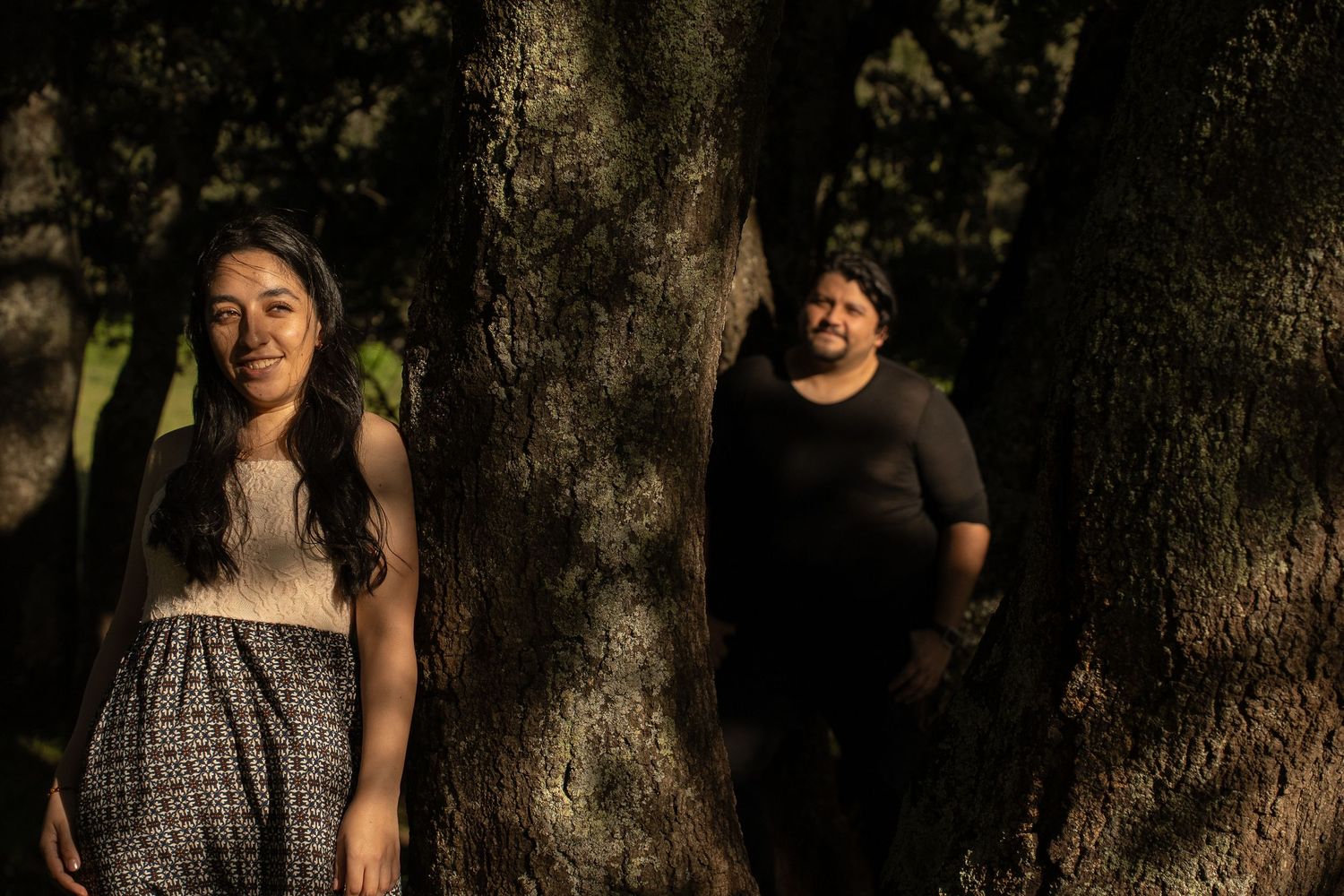 A couple stands between large tree trunks in dappled forest lighting.