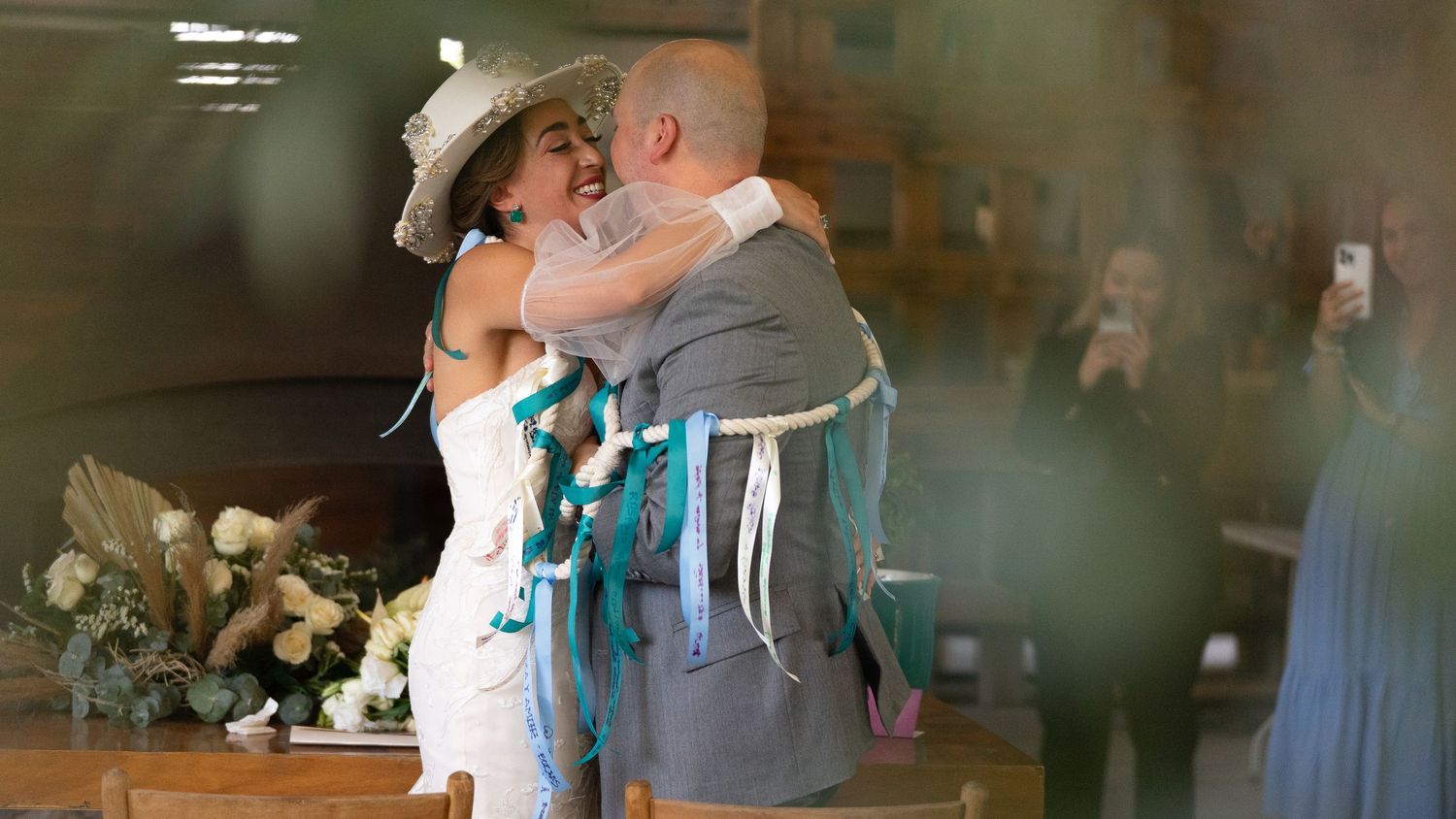 Bride and groom share first kiss at rustic wedding ceremony, bride wearing white dress and cowboy hat with teal ribbons.