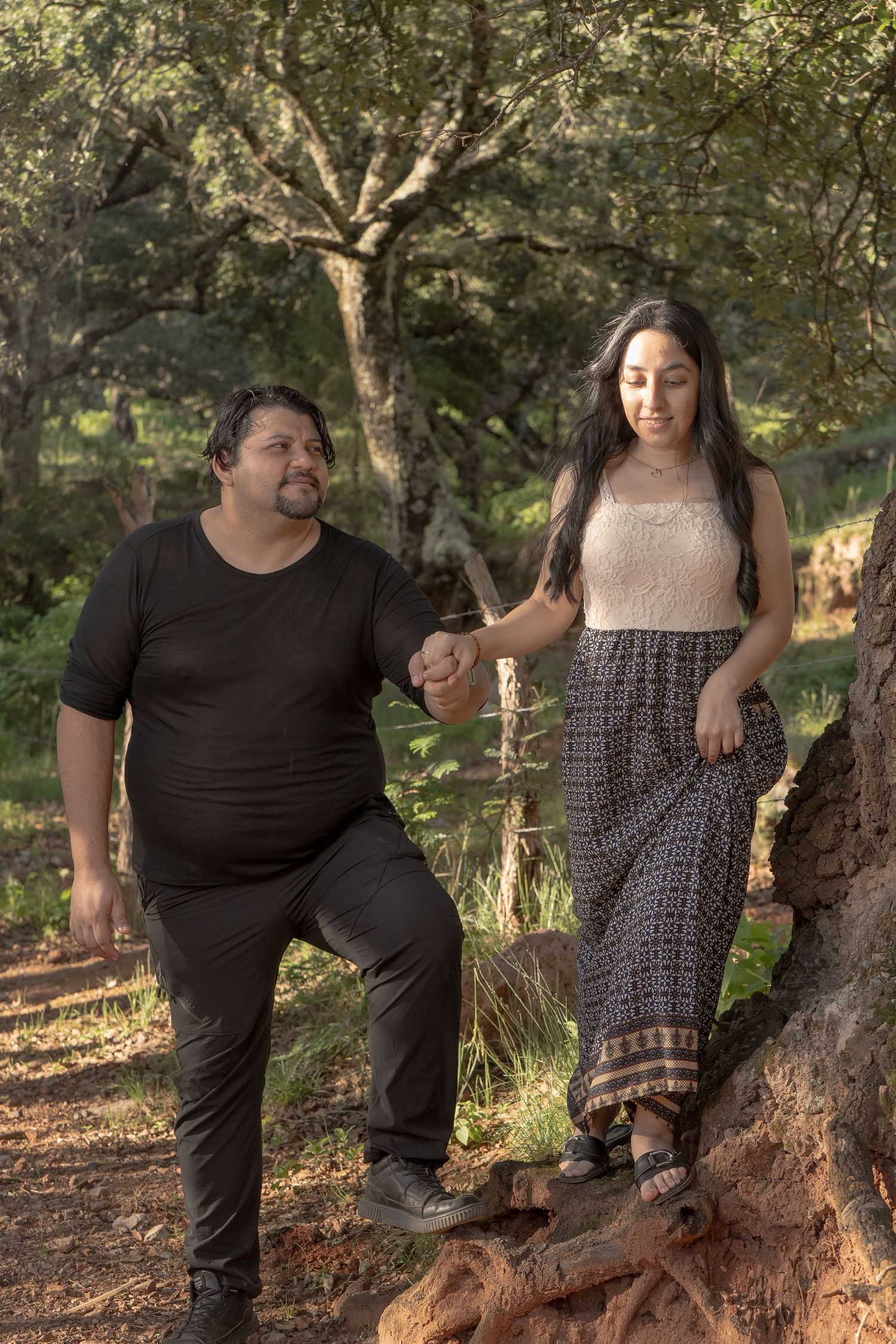 A couple stands together on tree roots in a wooded area during golden hour.