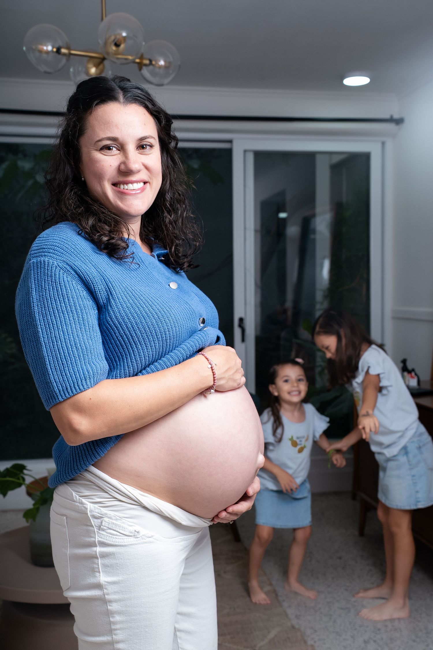 A pregnant woman in a blue sweater and white pants poses in a modern living room.