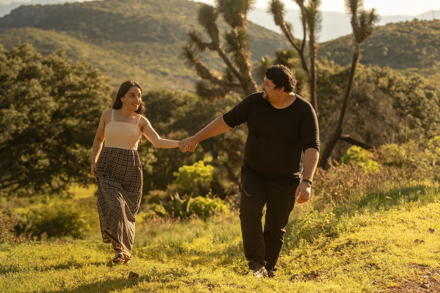 A couple walks hand in hand through a scenic mountain landscape at golden hour.