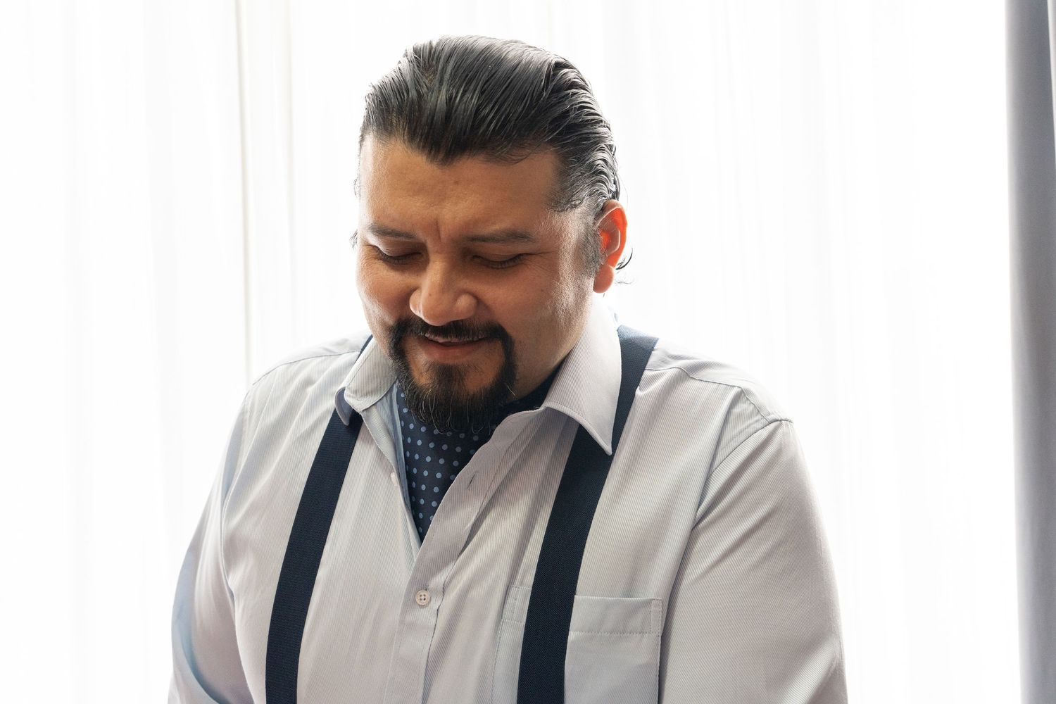 Professional in white dress shirt and suspenders smiles while working at desk with laptop and documents.