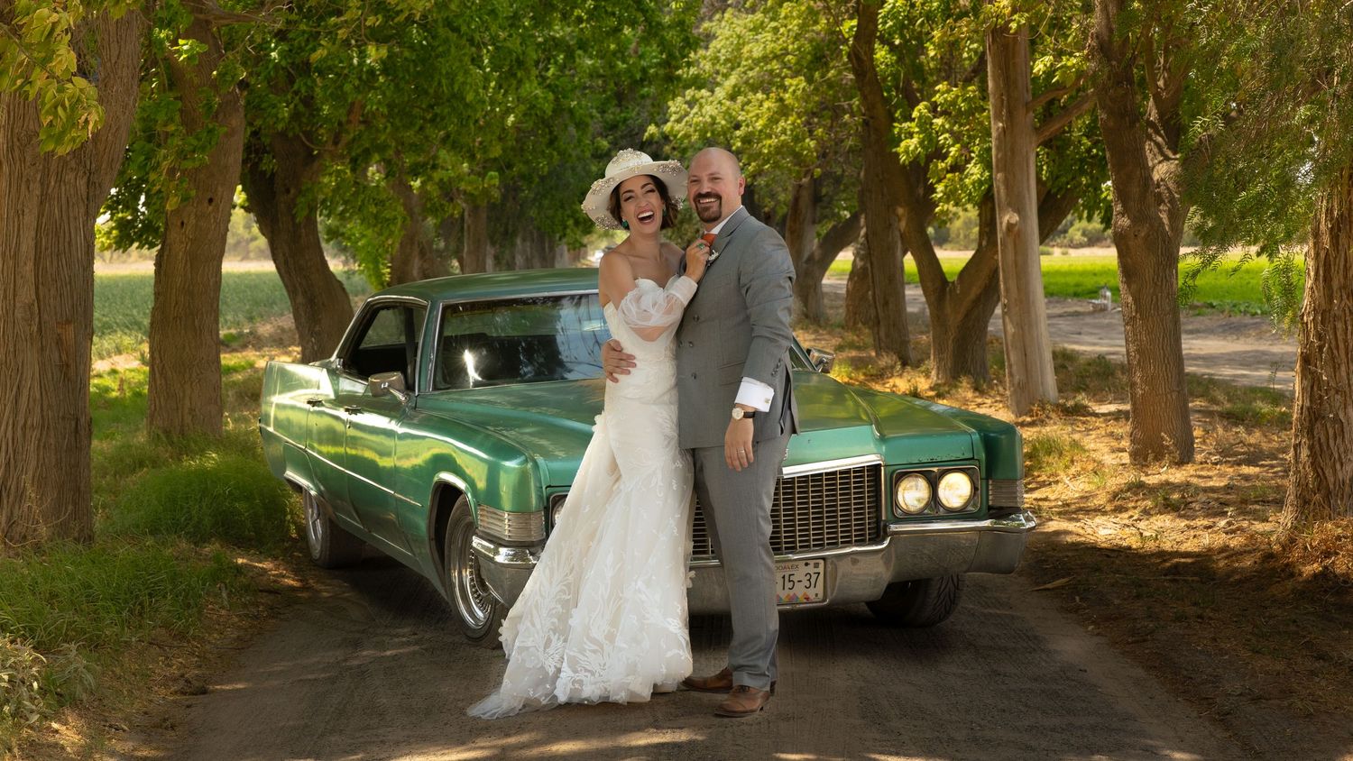 Bride and groom posing beside a vintage green Cadillac on a tree-lined dirt road on their wedding day.