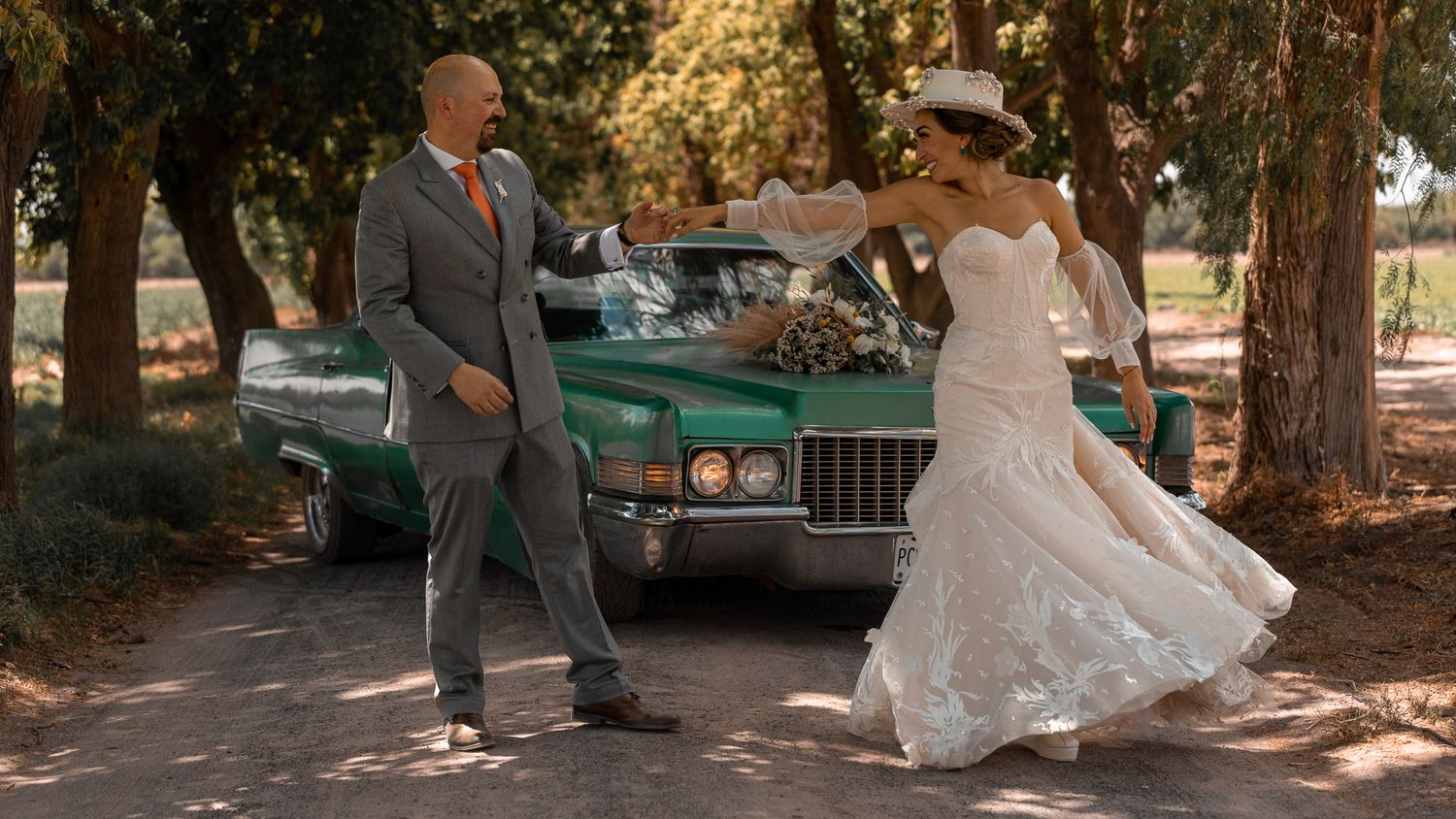 A couple dances joyfully on a tree-lined road with a vintage turquoise Cadillac in the background.