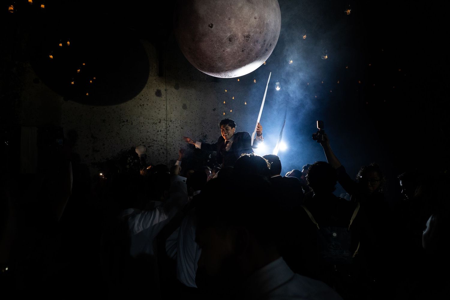 Dramatic stage performance with performer wielding sword under glowing moon backdrop surrounded by crowd.