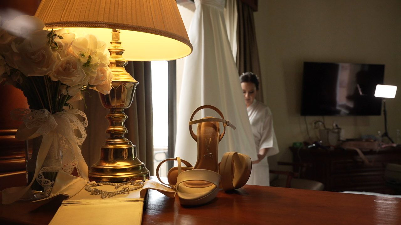 Elegant hotel room with brass lamp, gold heeled sandals on wooden table, and woman in white robe by curtained window.