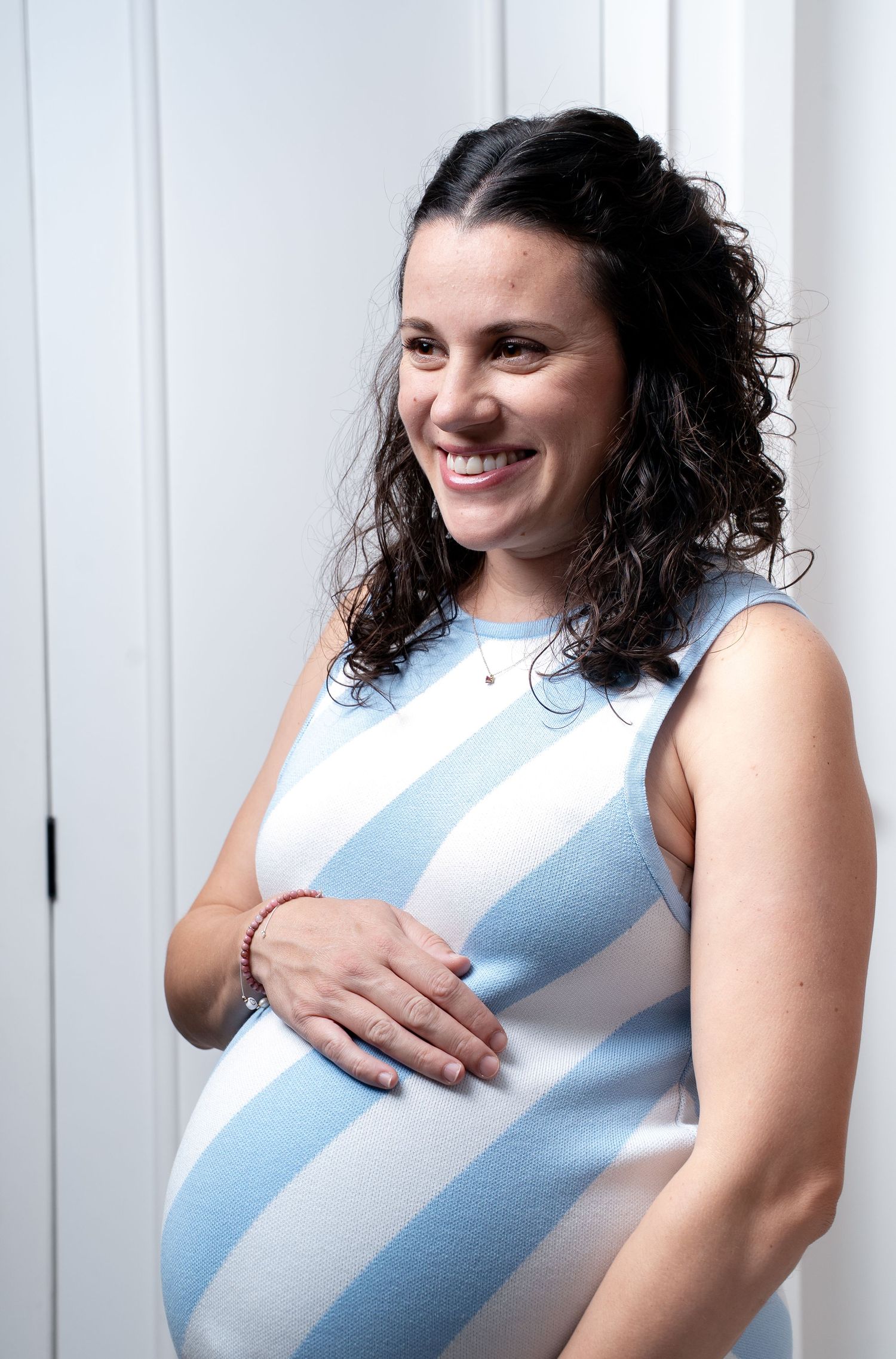 A pregnant woman in a blue and white striped dress smiles while touching her baby bump.