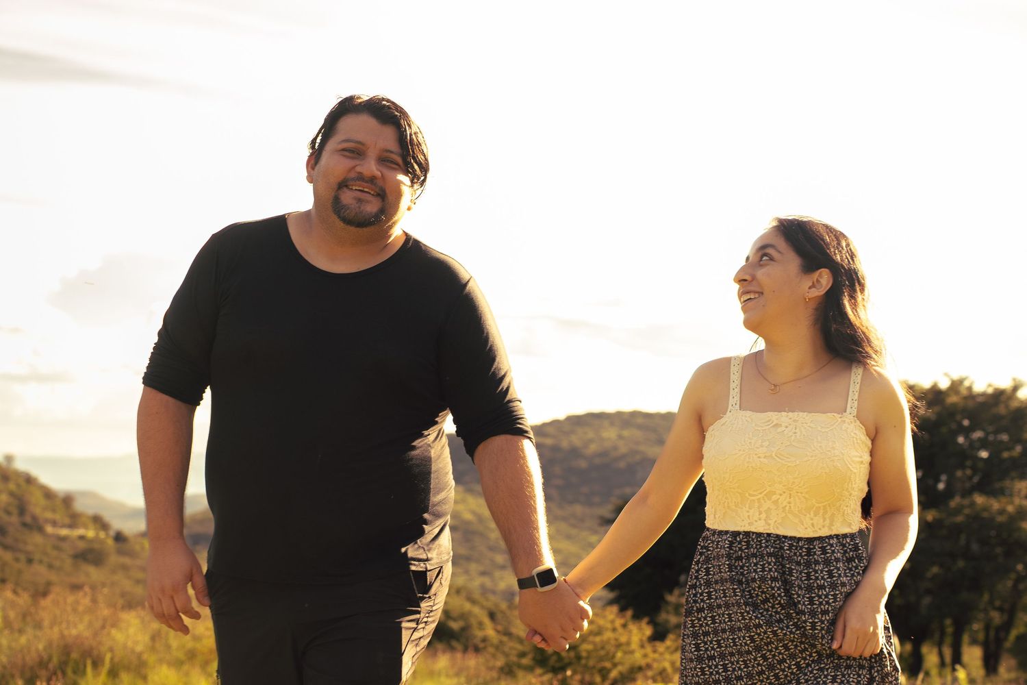 A happy couple holds hands while walking together at sunset on a hillside.