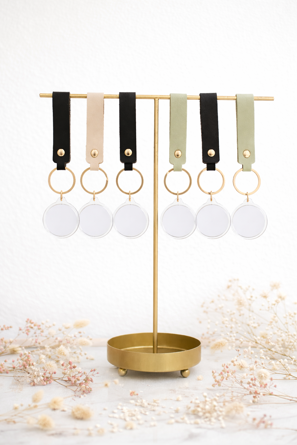 A gold display stand holds 6 pairs of keychains with round clear tags and leather straps in black, beige, and light green.