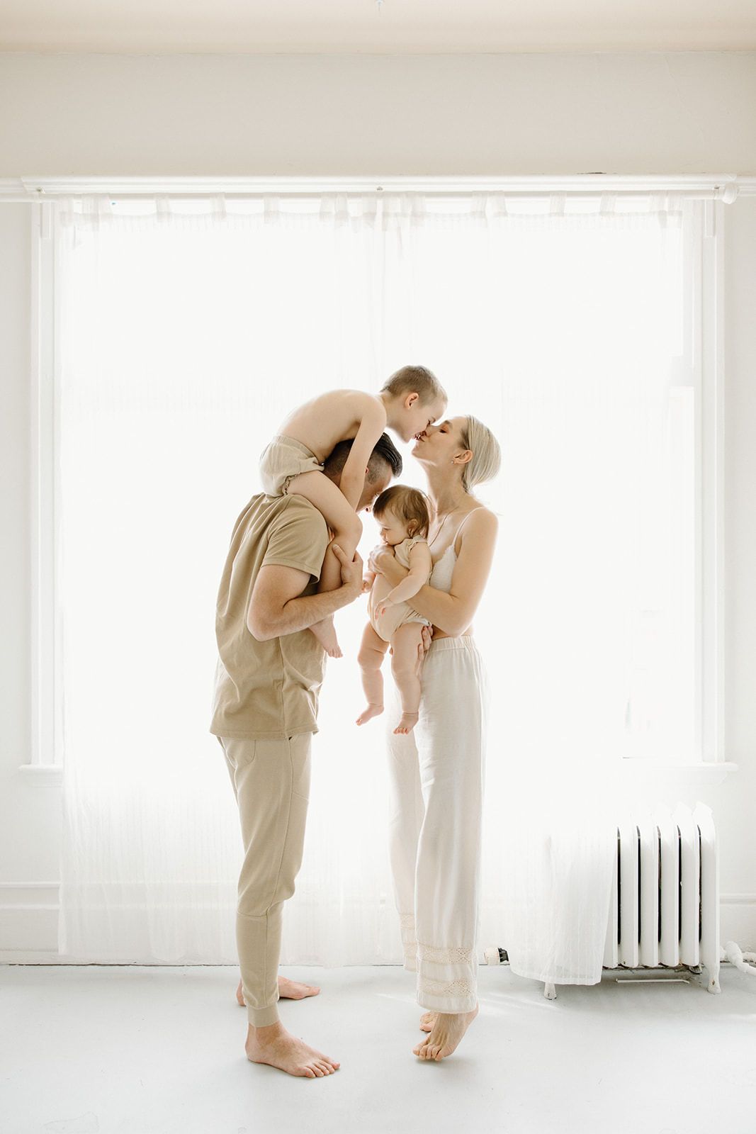 A family in neutral beige and white attire poses together against a bright window with a minimalist aesthetic.