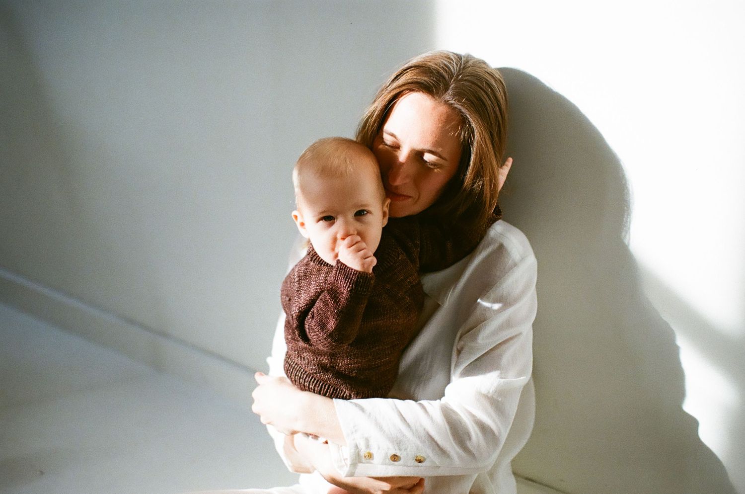 A mother in a white sweater tenderly holds her baby dressed in a brown knit outfit against a light gray wall.