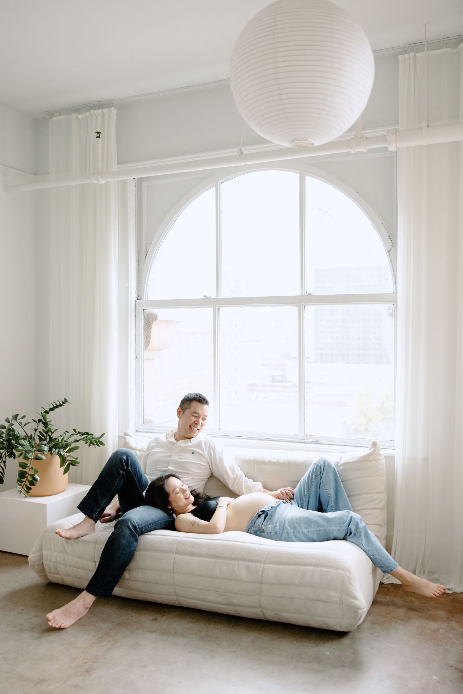 A couple relaxes together on a white couch near a large arched window with a paper lantern hanging above.