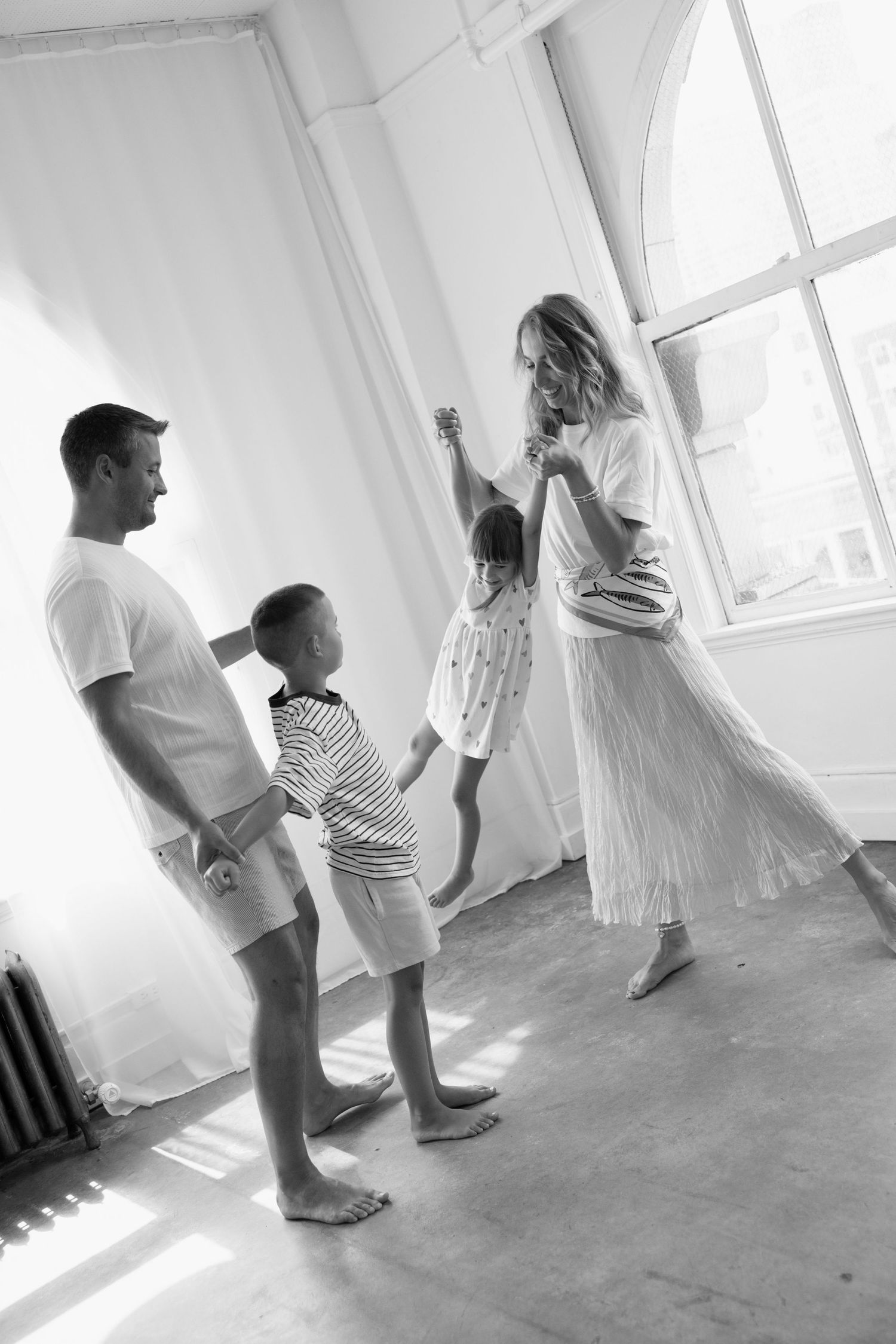 Black and white photo of a family of four playing in a bright, sunlit loft as parents swing a young child between them.