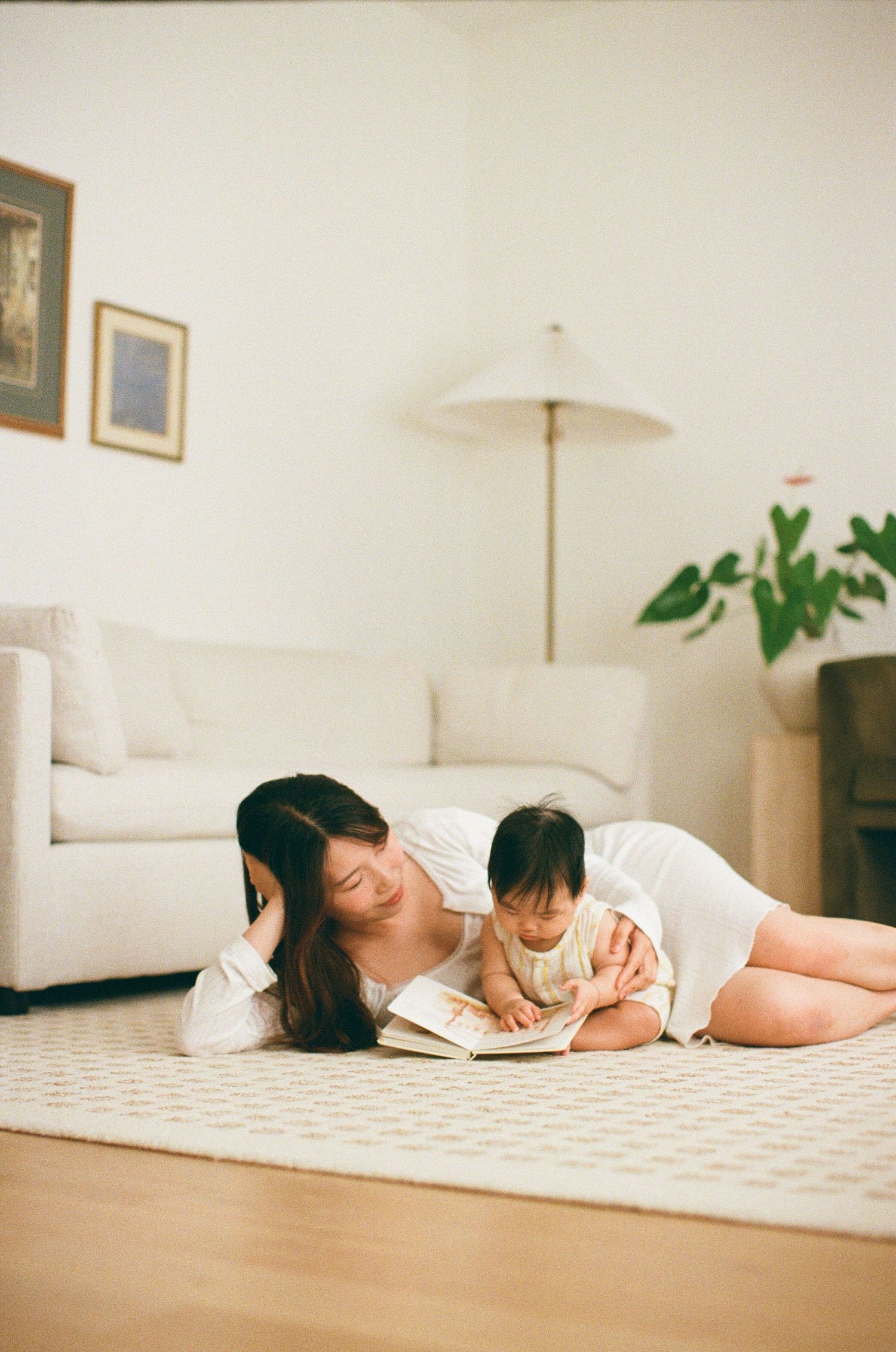 A mother lies on a carpet reading a book with her baby in a bright, minimalist living room.