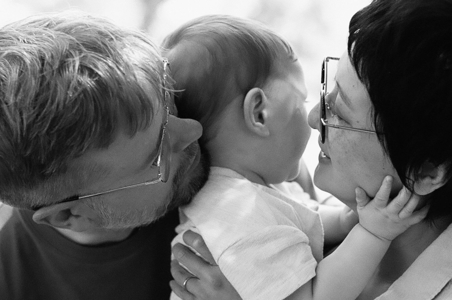 Black and white photo of grandparents lovingly kissing a smiling baby on both cheeks.