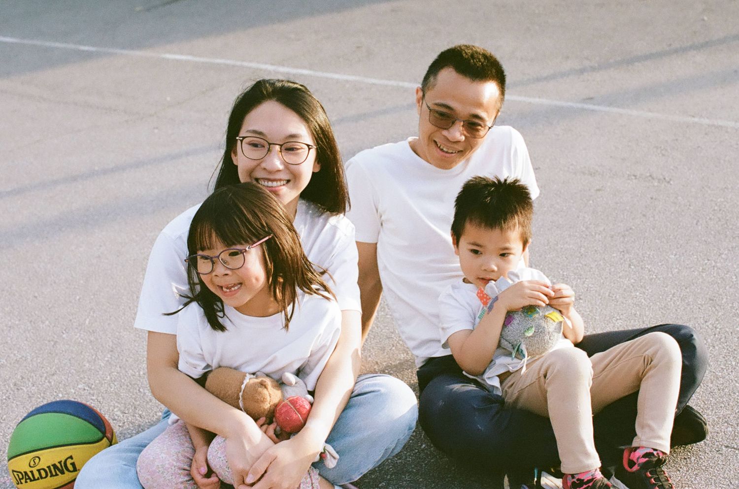 A happy Asian family of four in white shirts sits together on a basketball court, smiling warmly at the camera.