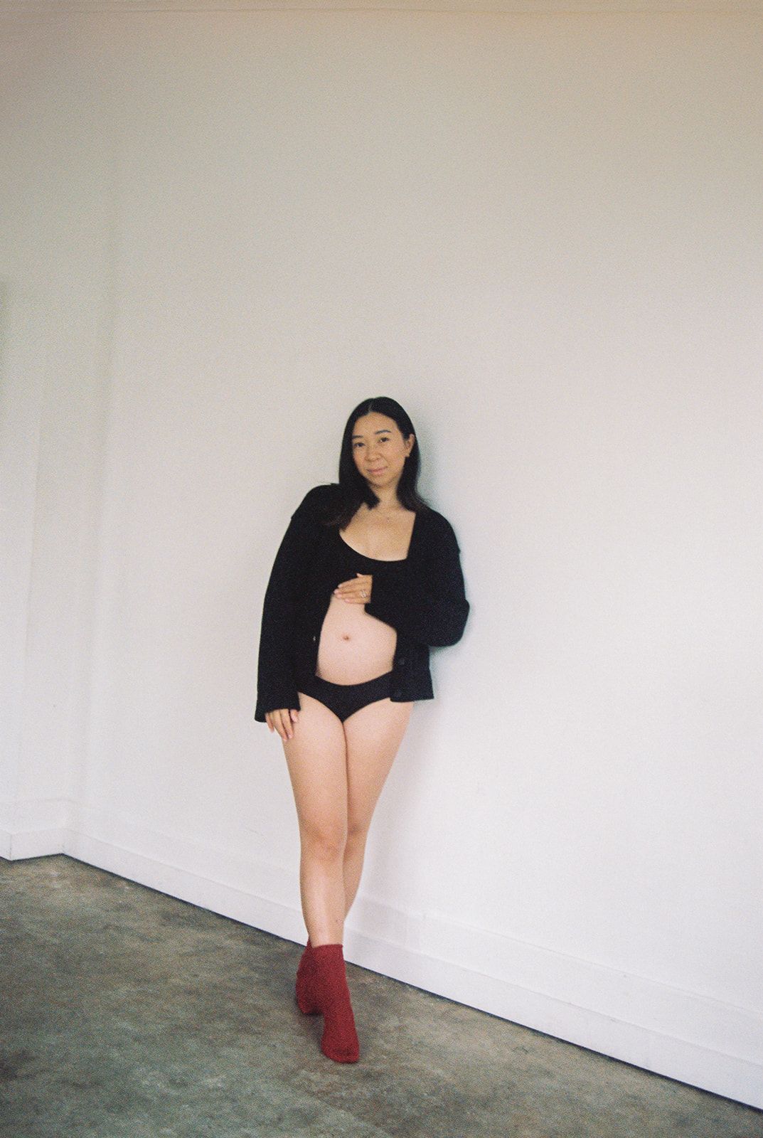 A person wearing a black cardigan over pink bodysuit and red ankle boots poses against a white wall.