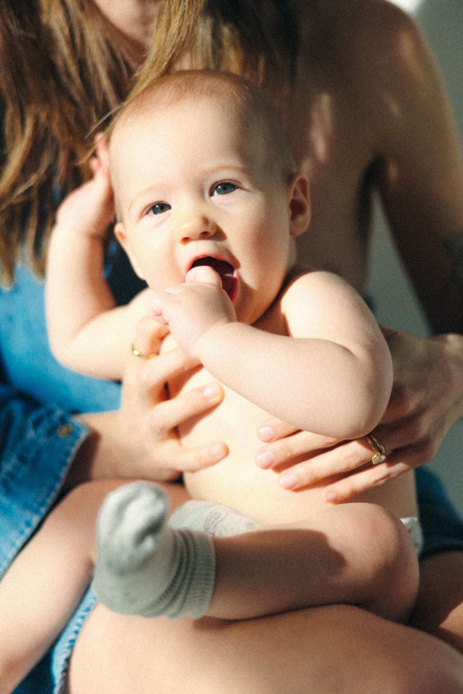 A happy baby in a diaper and grey socks being held by a woman in a blue denim top in natural light.