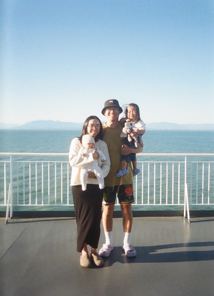 A family stands together on a ferry deck against a scenic ocean and mountain backdrop on a clear sunny day.
