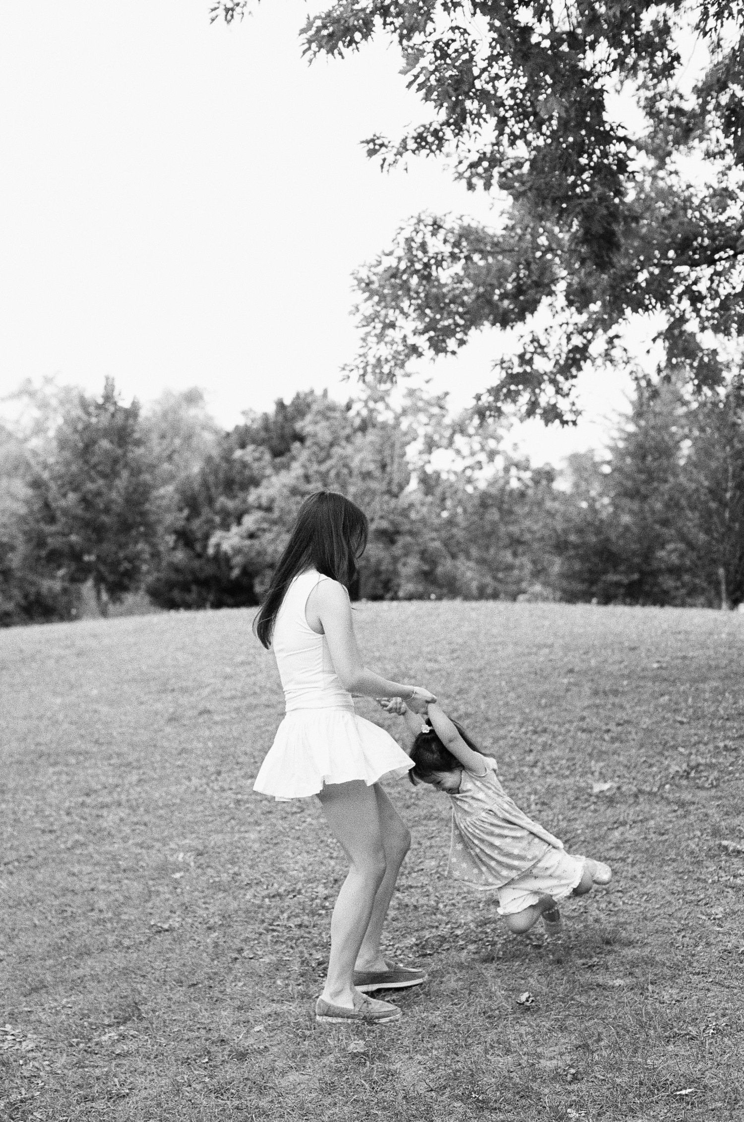Black and white photo of a woman spinning a young child by the hands on a grassy park lawn with trees in background.