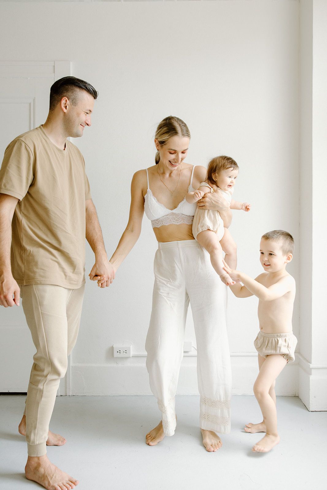 A family dressed in neutral tones holds hands while standing together against a white wall.