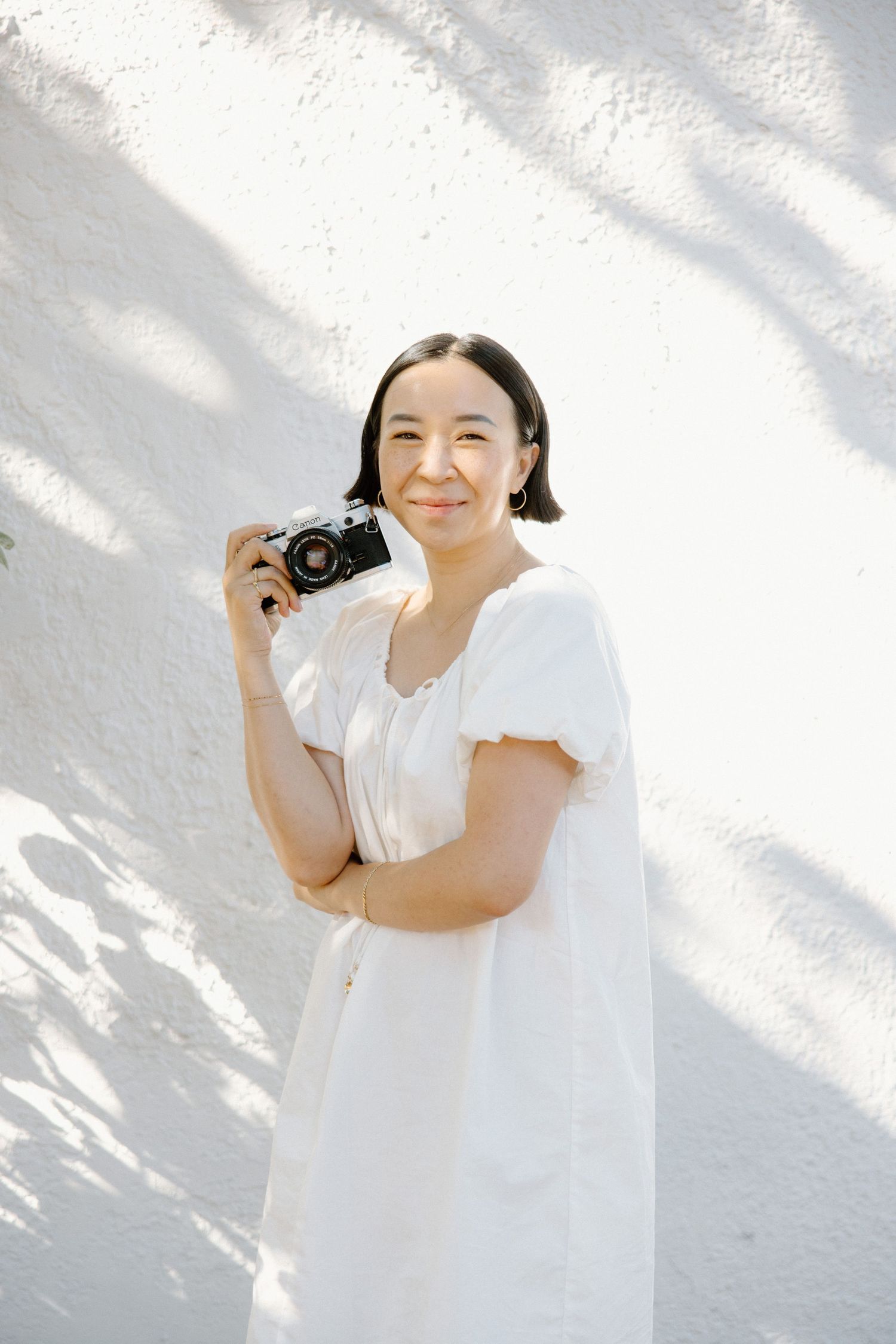 A photographer in a white summer dress holds a vintage camera against a bright white wall with dappled shadows.