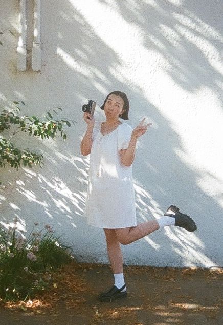 A person in a white dress strikes a playful pose while holding a camera against a sunlit wall with leafy shadows.