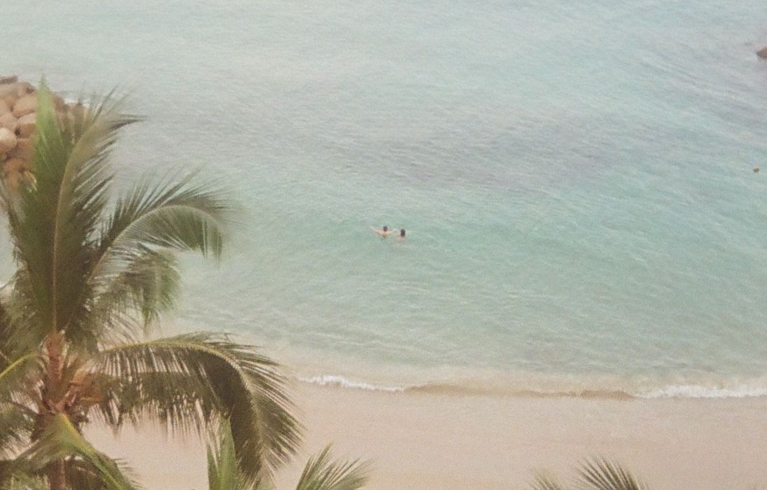 Aerial view of a tropical beach with turquoise water, palm trees, and distant swimmers enjoying the ocean.