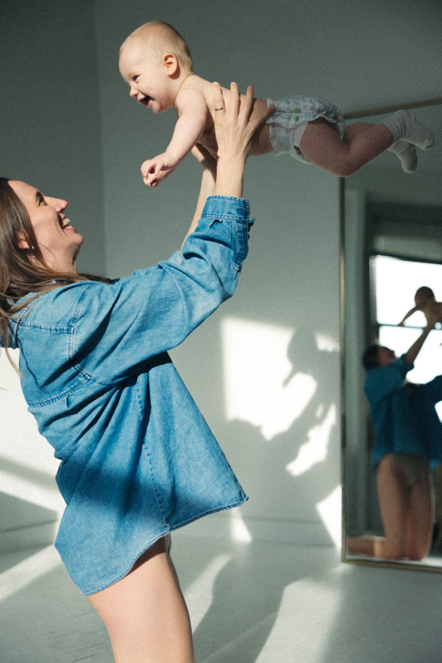 A joyful mother in a denim shirt lifts her laughing baby overhead in a bright white room with a mirror reflection.