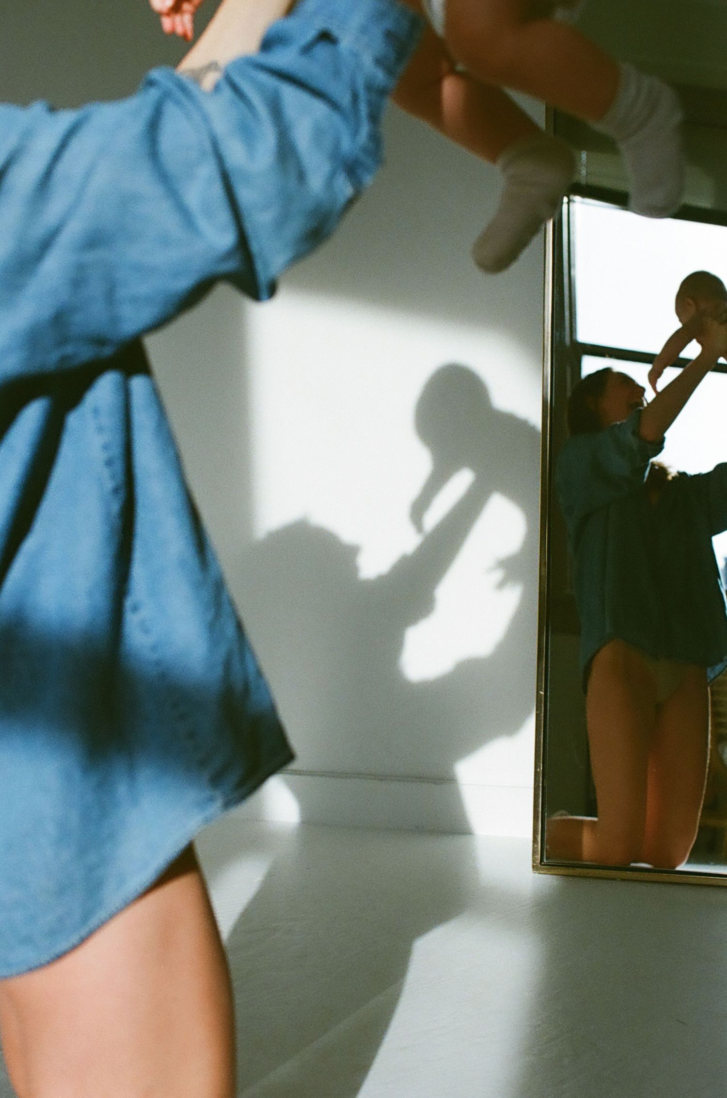 Person in blue denim shirt reaching up, casting dramatic shadow on white wall beside a mirror reflection.