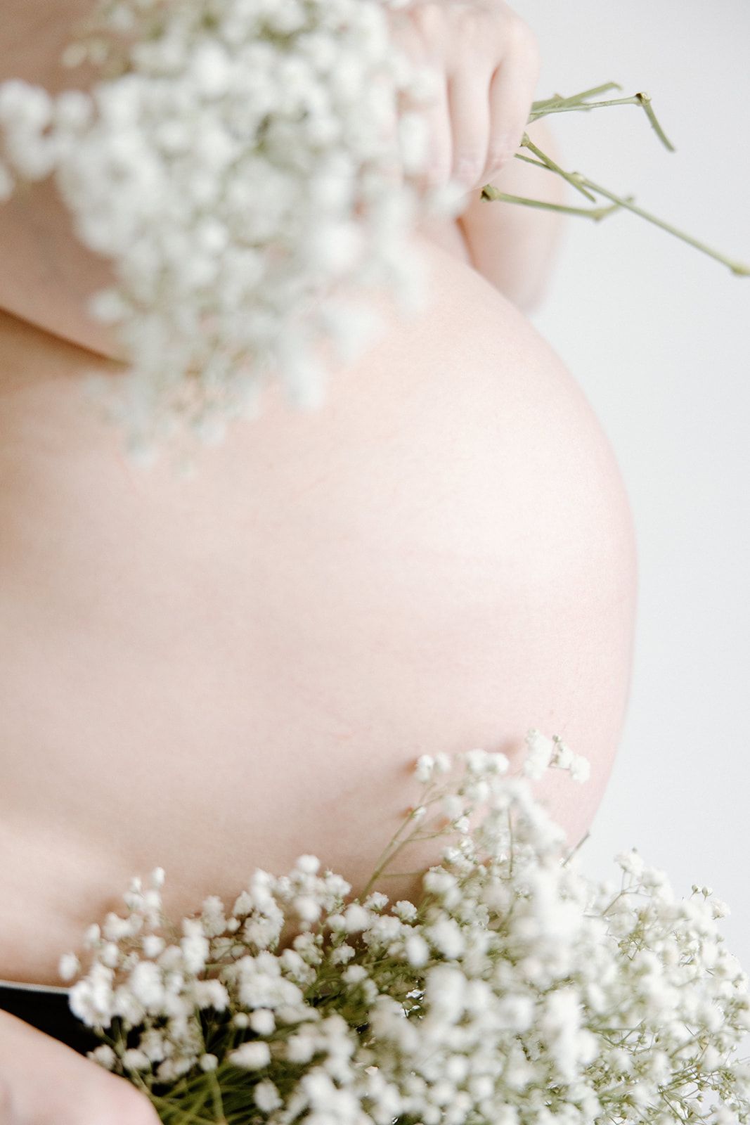 Delicate white baby's breath flowers frame a pregnant woman's bare belly in a soft, artistic maternity photo.