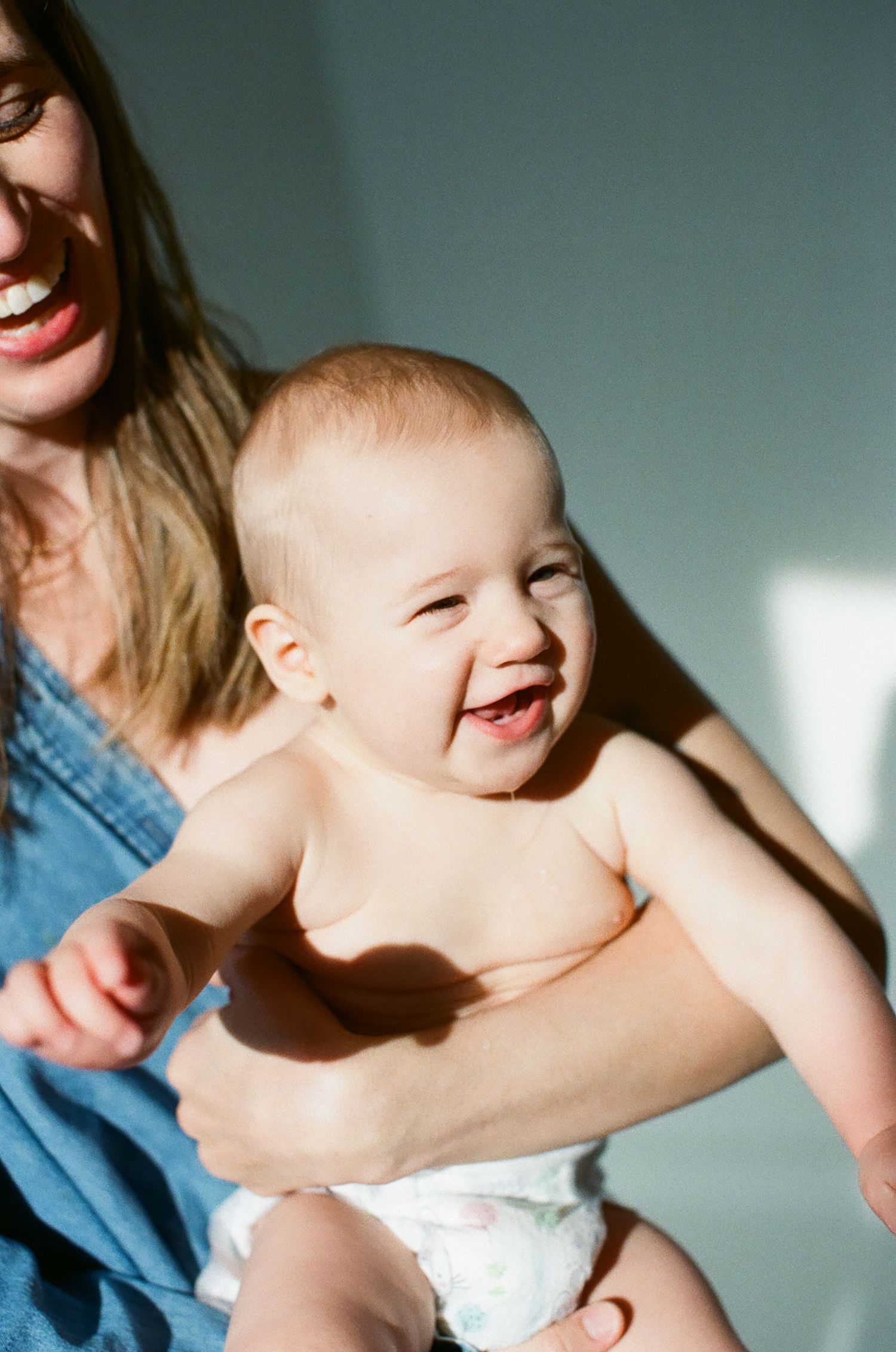 A joyful baby in a diaper laughs while being held by a smiling woman in a blue denim top.
