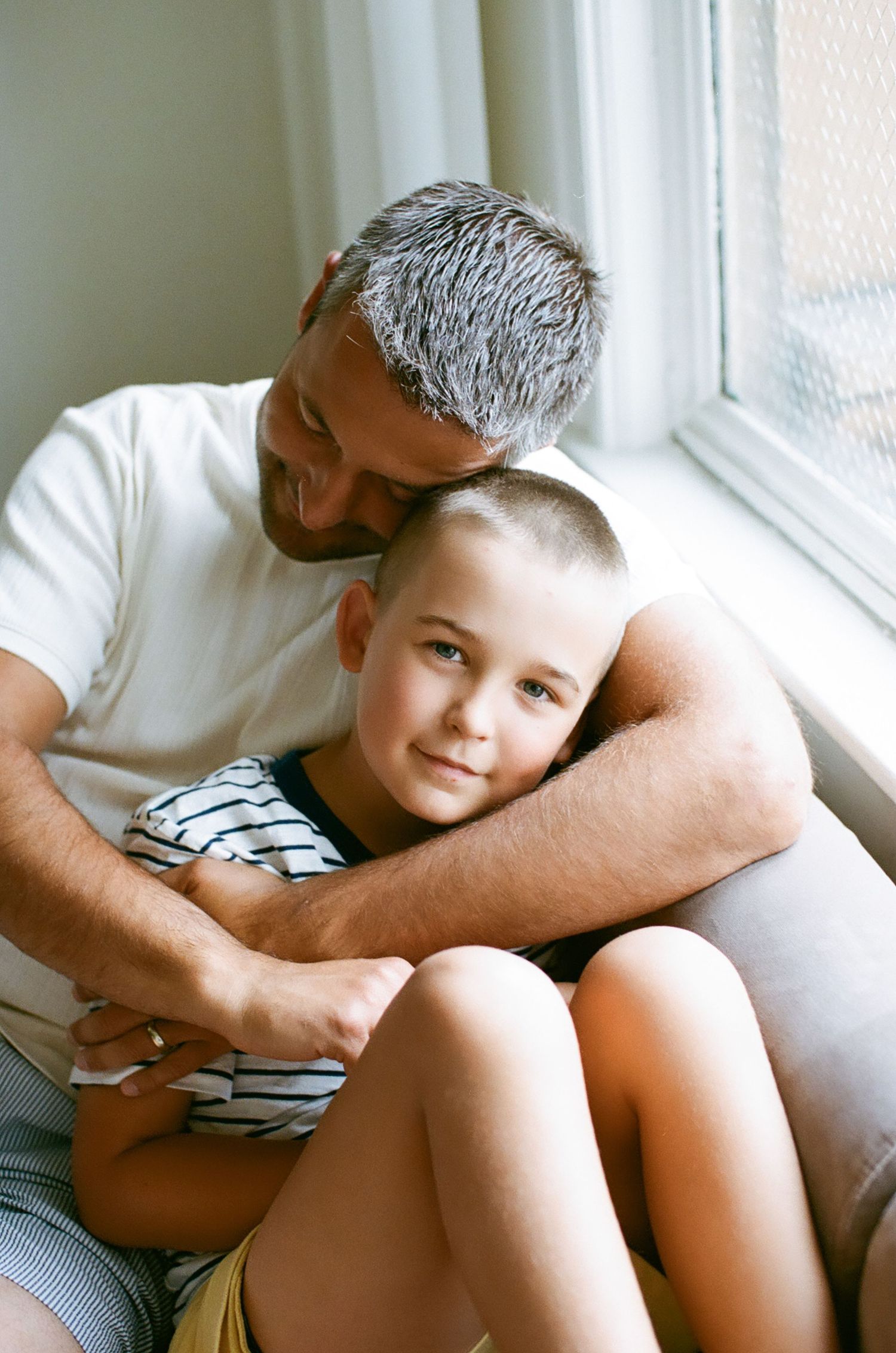 A father with gray hair hugs his young son tenderly while sitting together by a sunlit window indoors.