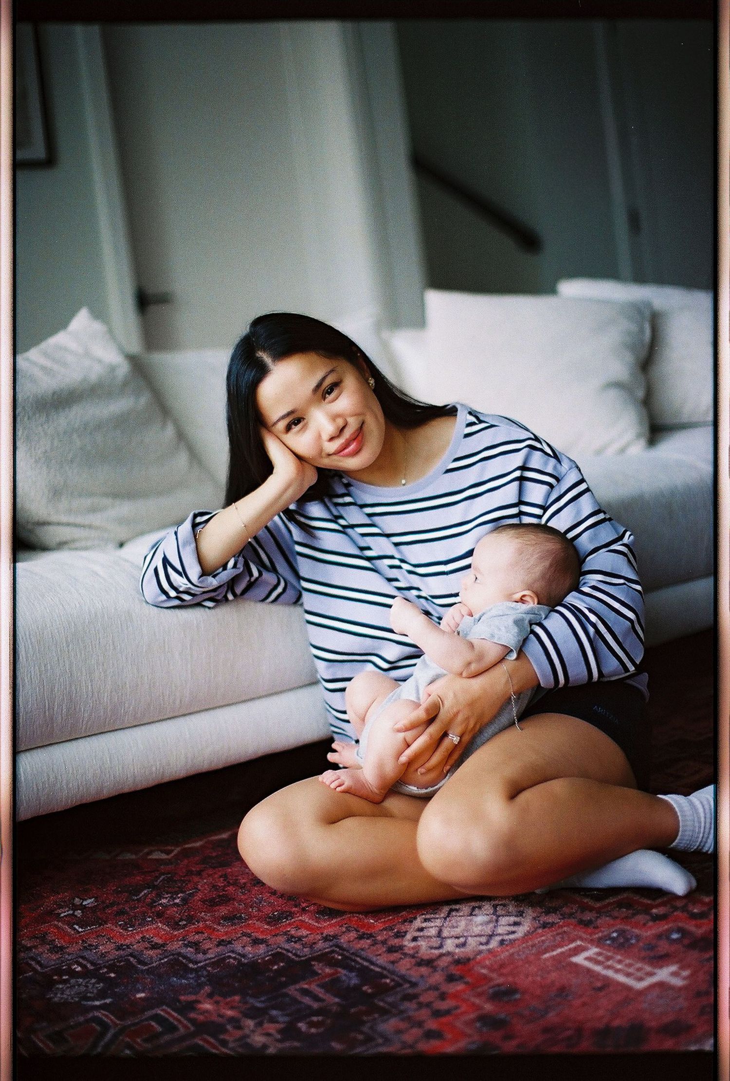 Young mother sitting cross-legged on a rug, cradling her newborn baby while smiling warmly in a cozy living room.