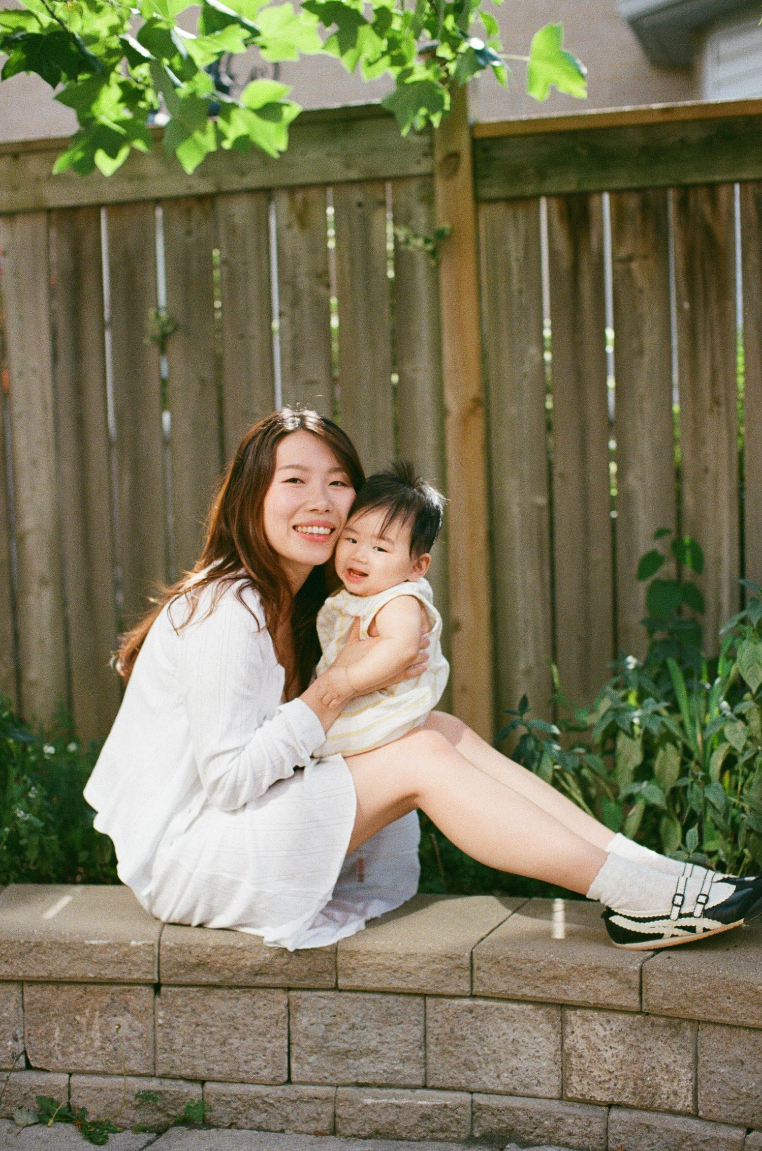 A smiling Asian woman in a white dress sits outdoors holding a baby in a yellow outfit near a wooden fence.