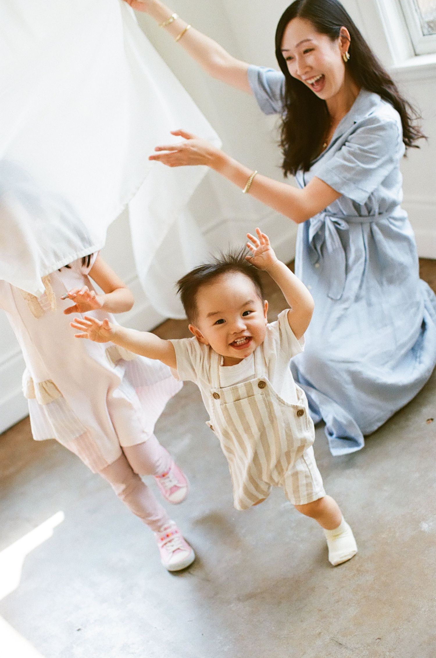 A smiling mother in a blue dress plays with two toddlers under a white sheet in a bright room.