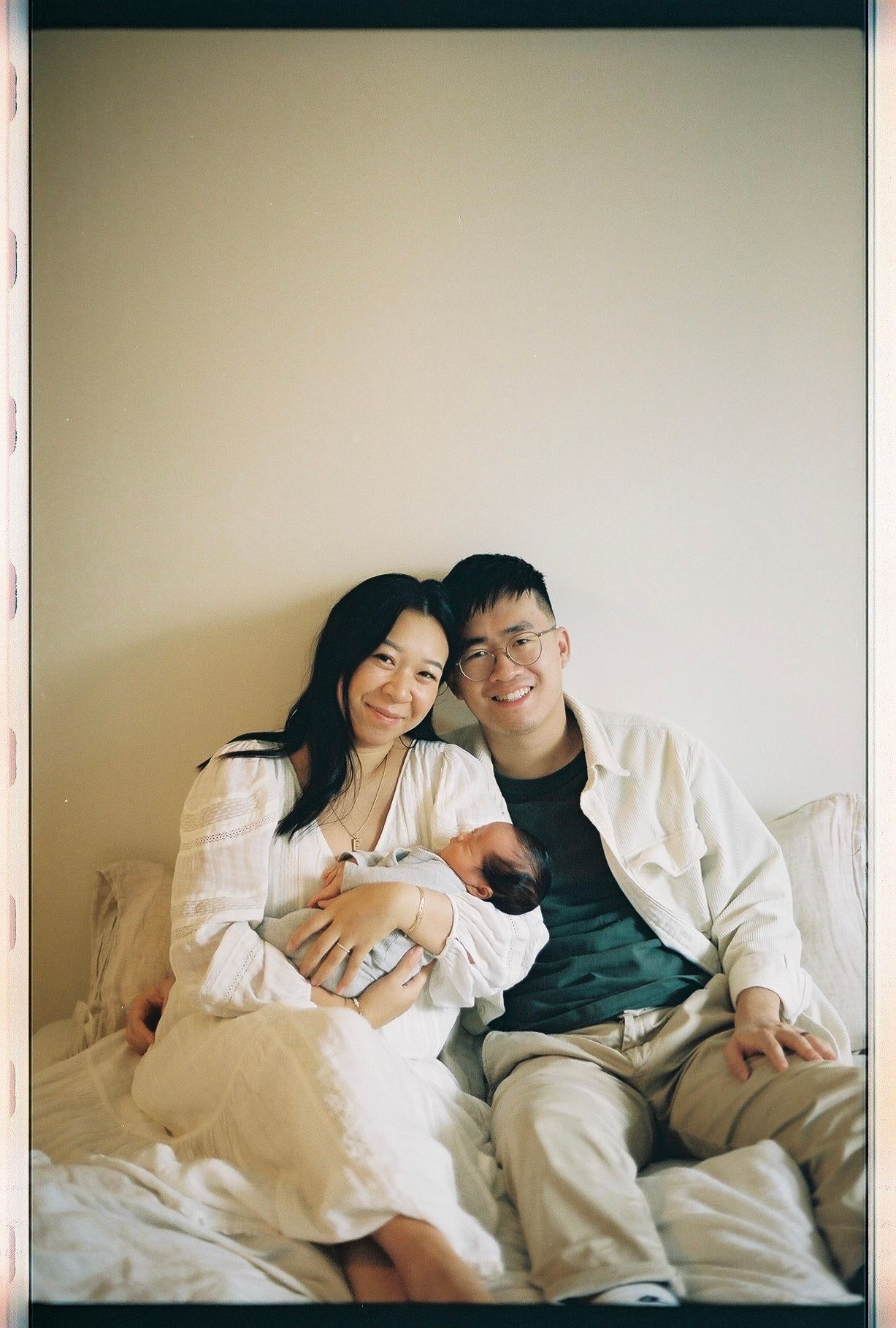 Young couple sitting together on bed with their newborn baby in a bright, minimalist setting.