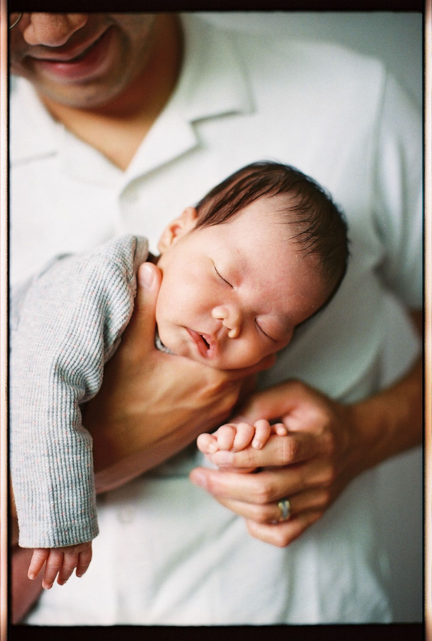 Newborn baby sleeping peacefully in father's arms, gently cradling tiny hands, wearing gray onesie.