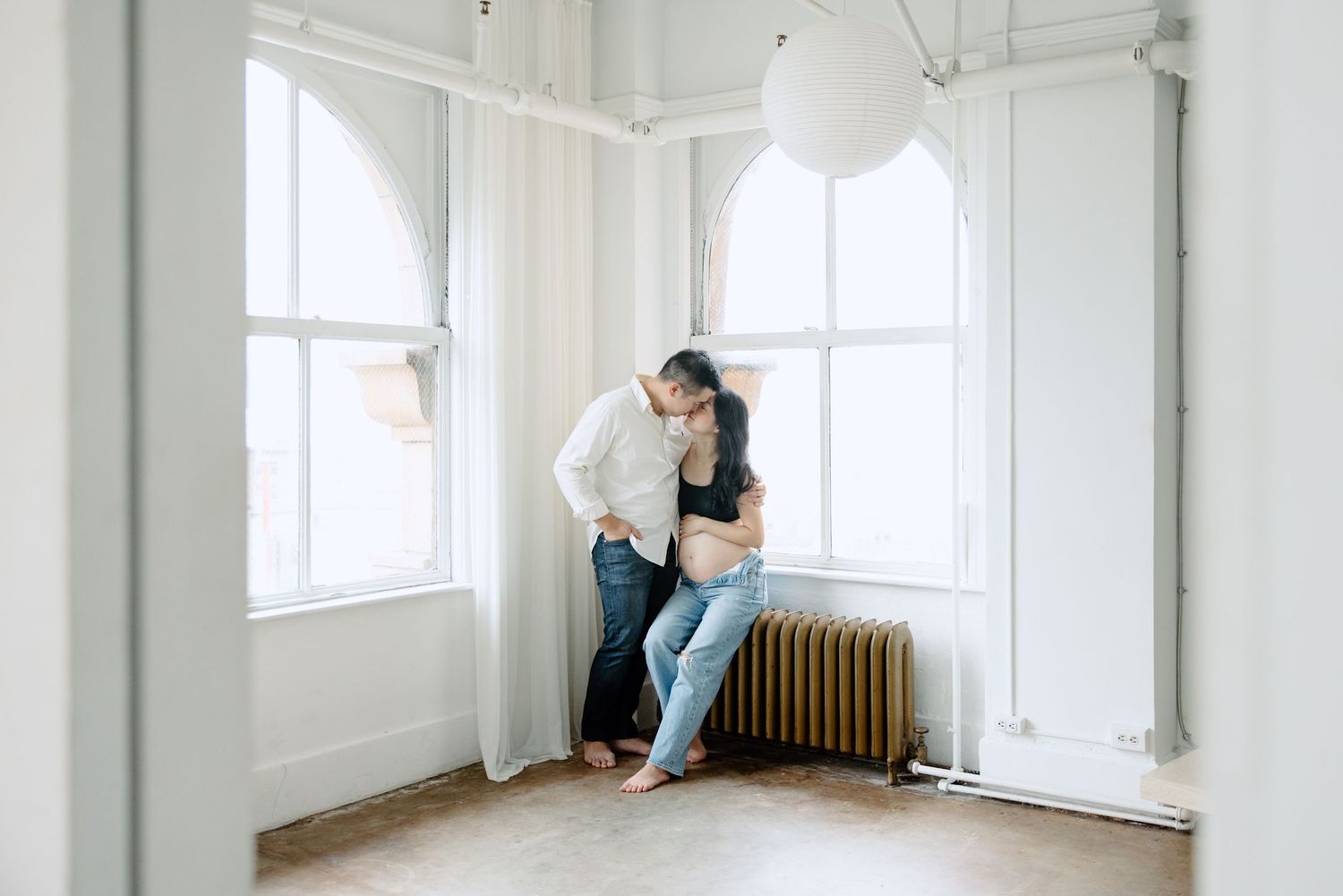 A couple embraces near a bright window in a minimalist white room with hardwood floors and a vintage radiator.