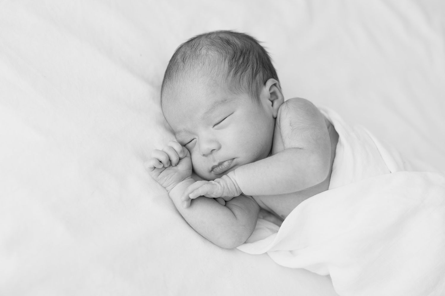 A peaceful sleeping newborn baby wrapped in white blankets, photographed in black and white.
