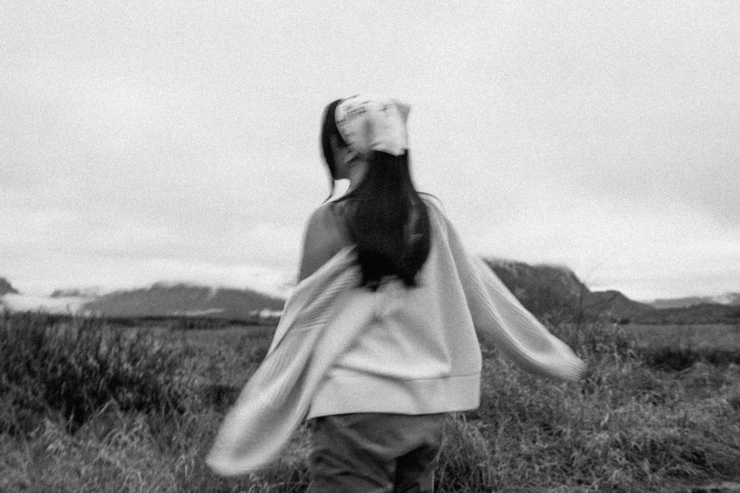 Black and white image of person twirling in flowing dress outdoors in natural landscape against cloudy sky.