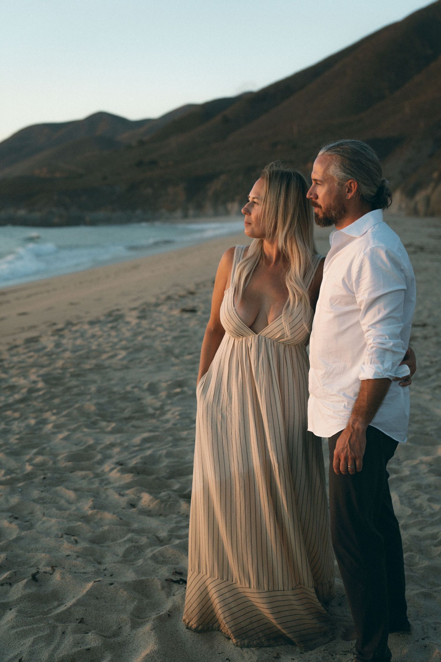 Couple in white and striped dress sharing intimate moments on a beach with mountains in background.