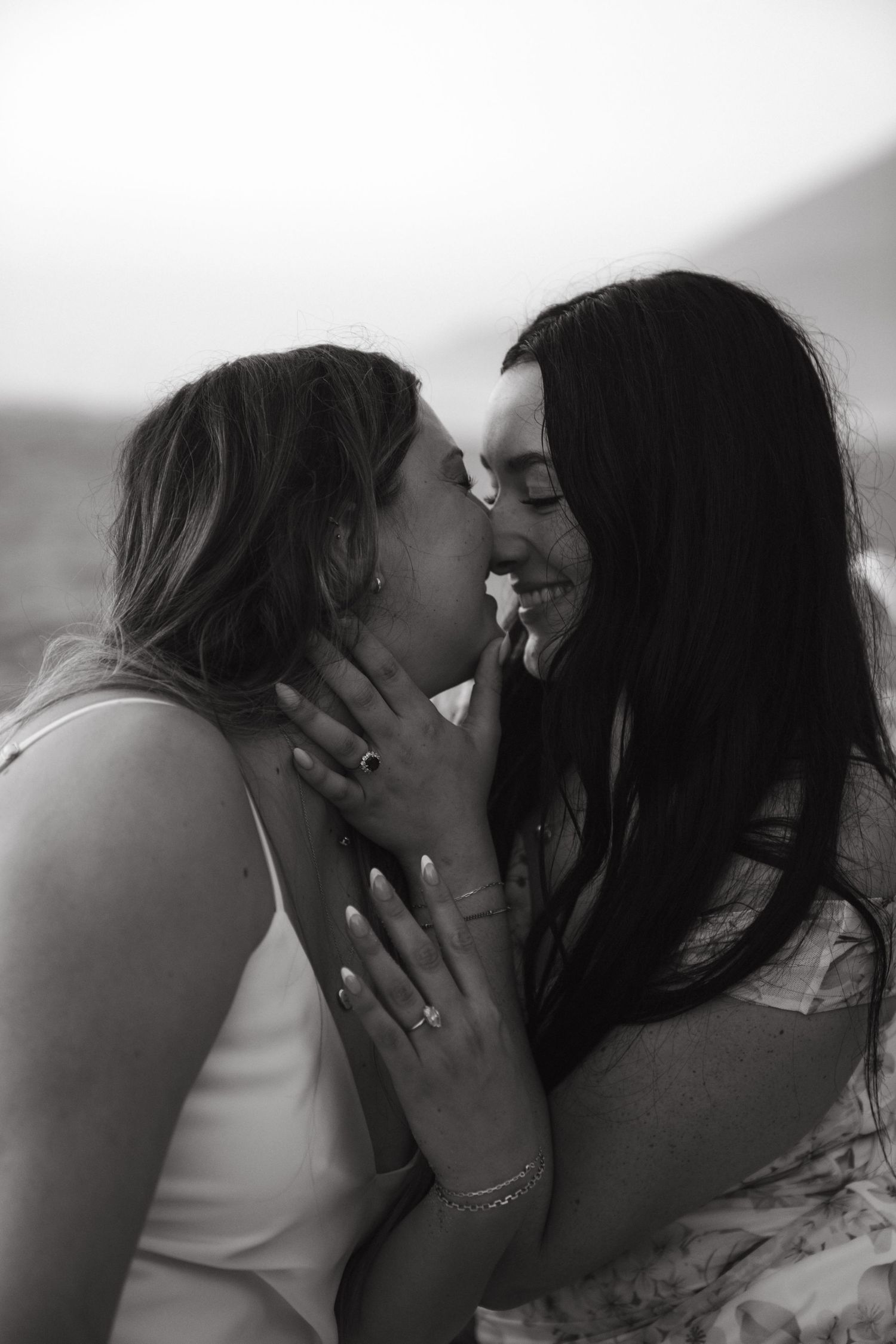 Black and white photo of two women sharing a tender moment, smiling and about to kiss, wearing rings and jewelry.